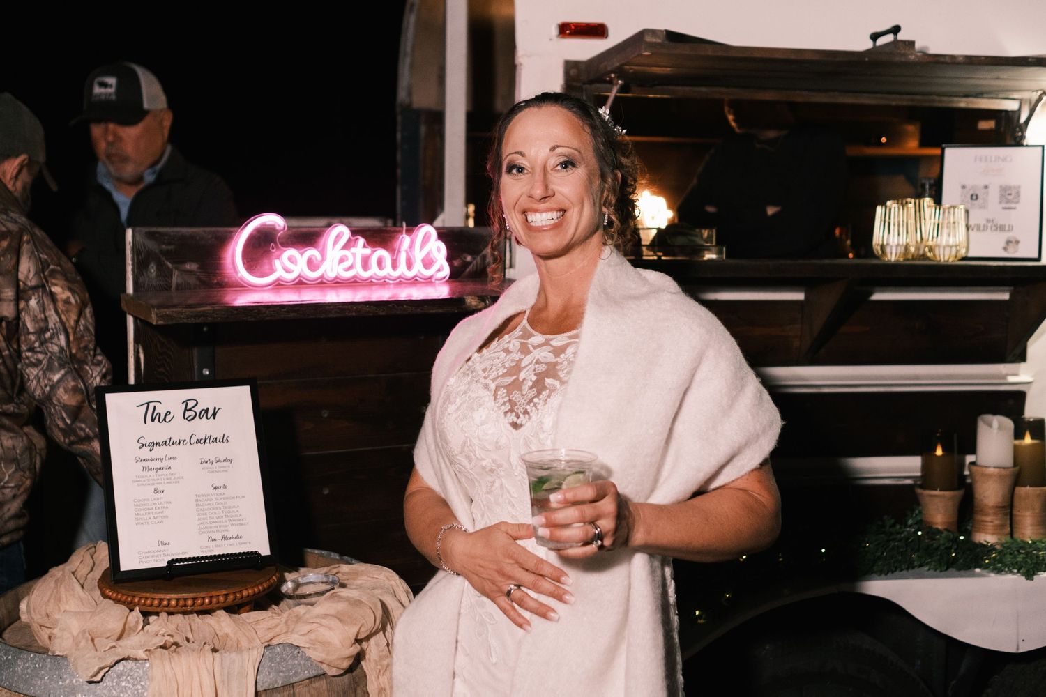 Smiling bride in white dress and fur wrap posing near a cocktails neon sign at reception.