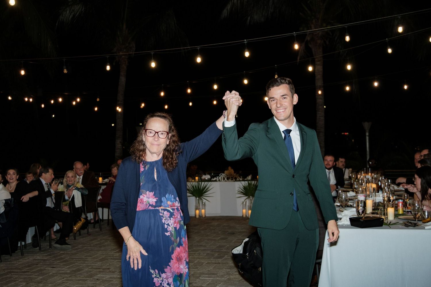 String lights illuminate an outdoor evening celebration with guests mingling.