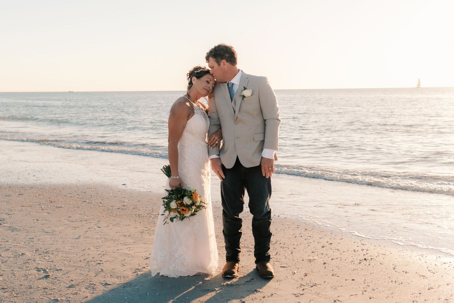 Newlyweds embrace on beach at golden hour, bride holding colorful bouquet by ocean.