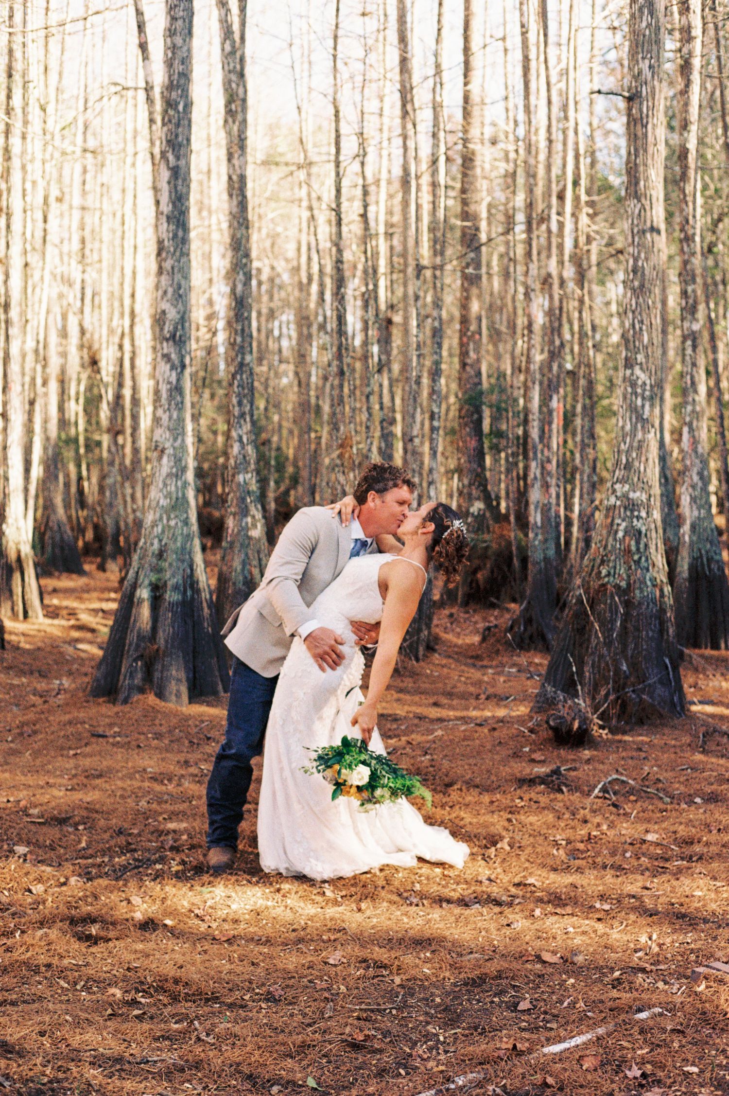 Groom dips bride for a romantic kiss among tall trees in a sunlit forest wedding photo.