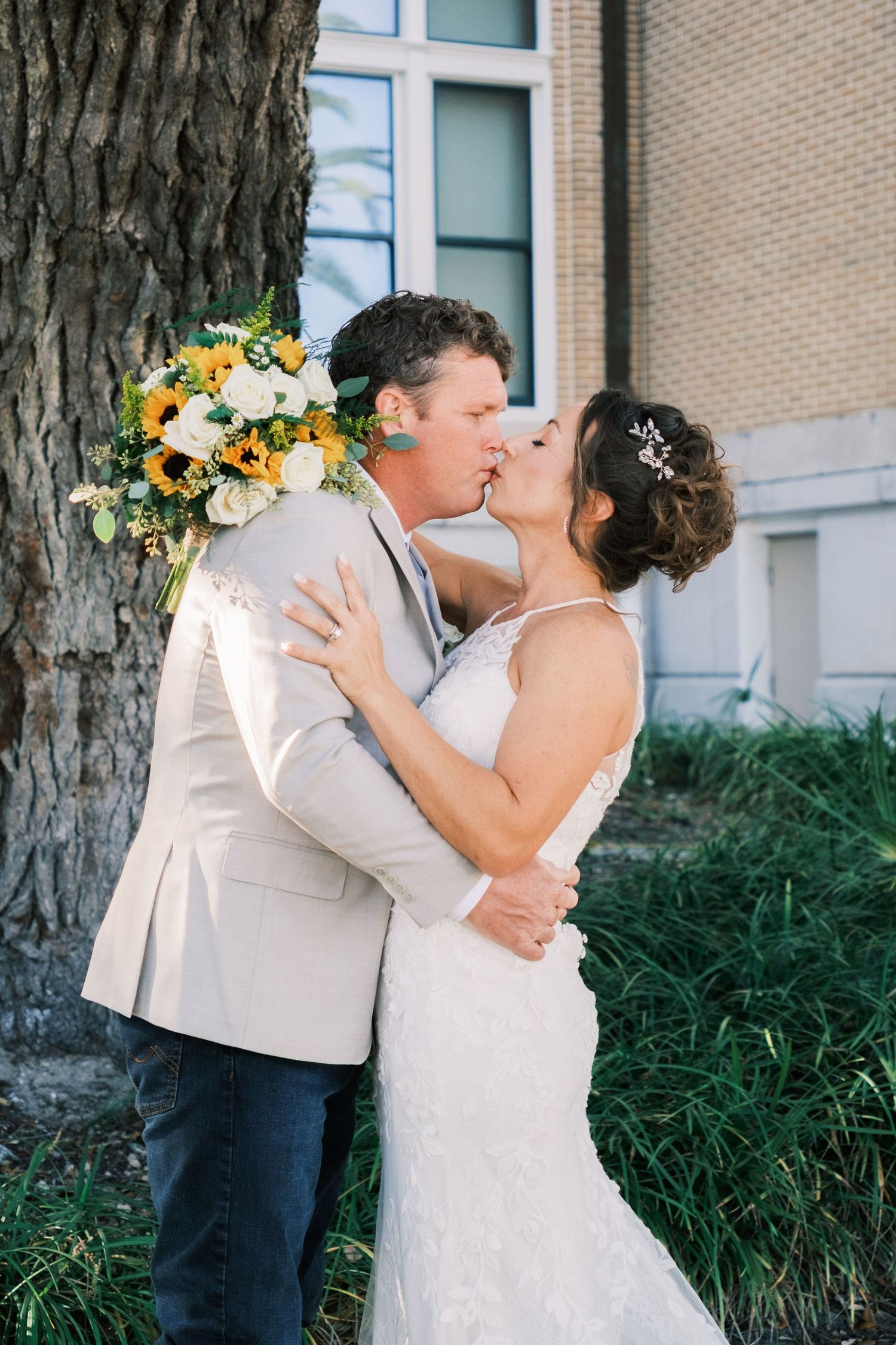 Bride and groom share a romantic kiss near a tree, bride holding a sunflower bouquet in a lace wedding dress.