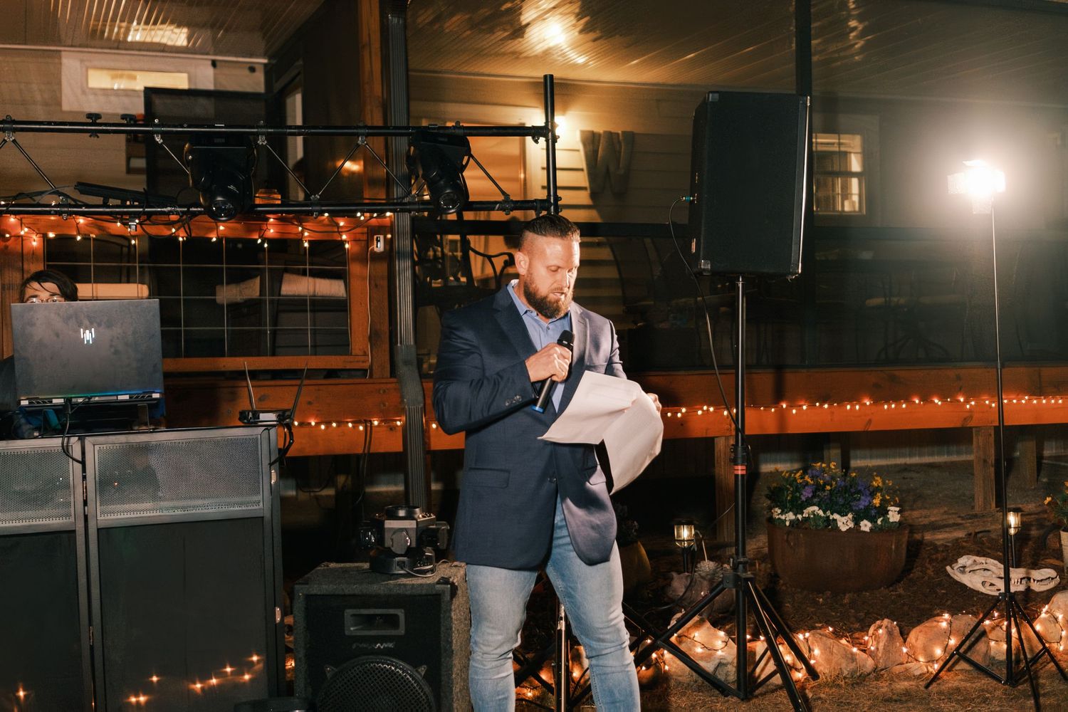 Man in navy blazer delivers speech with microphone at decorated wedding reception venue.