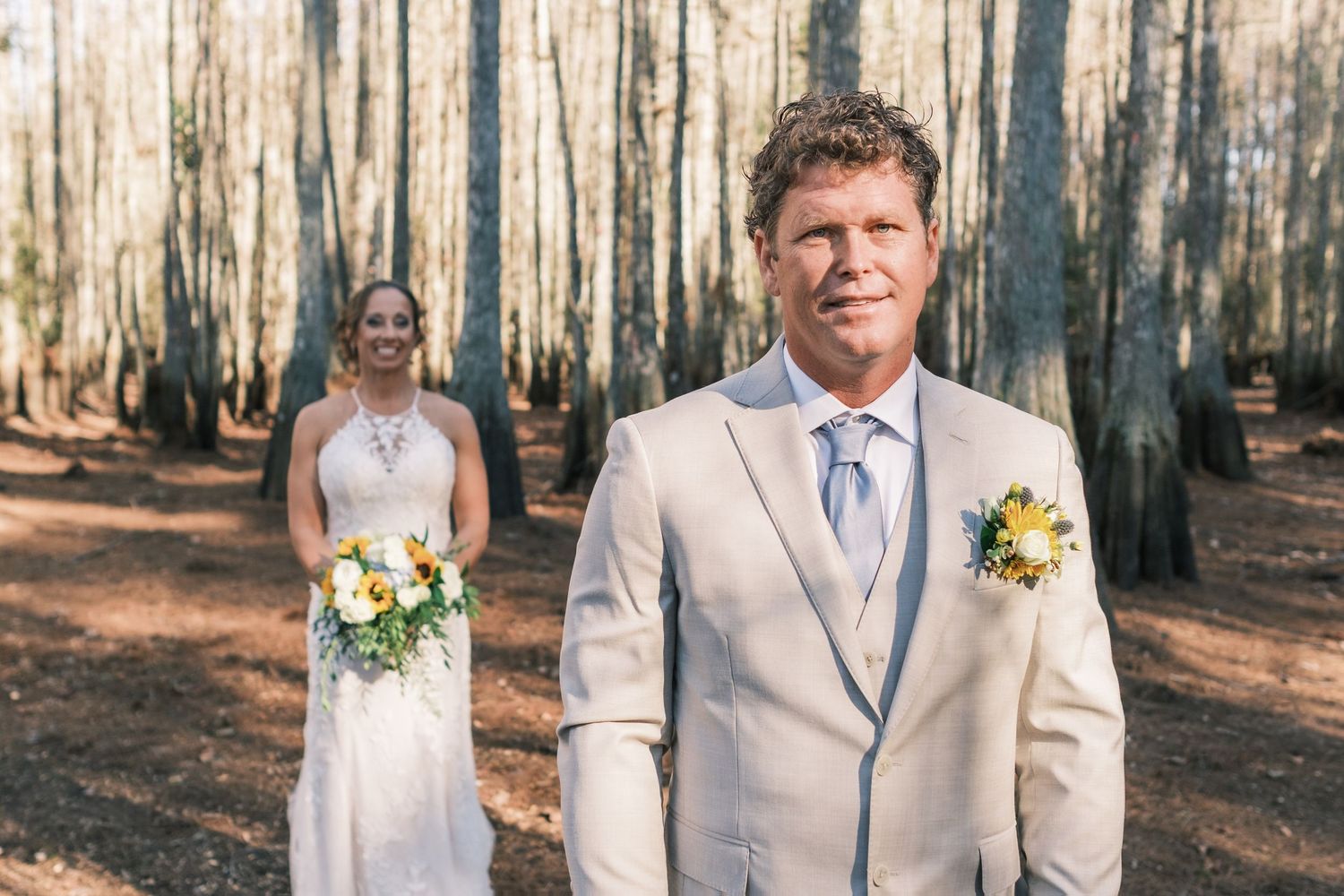Groom in tan suit stands before bride in cypress tree forest for first look.