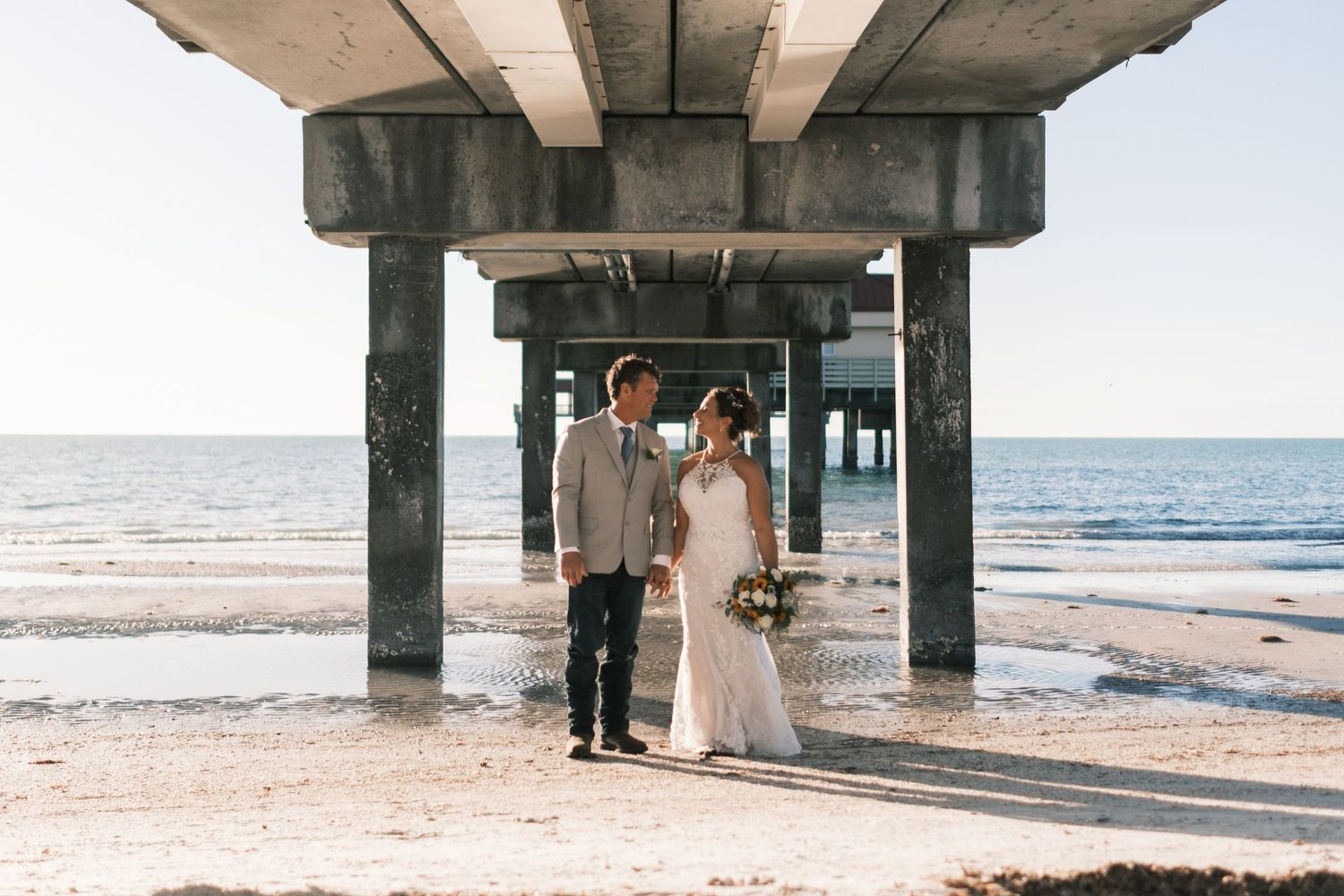 Bride and groom walk hand in hand under concrete pier on sandy beach.