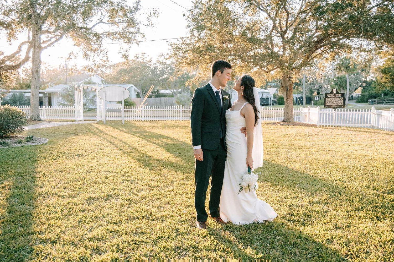 A wedding couple poses on a sunlit lawn with white picket fence and trees in background.