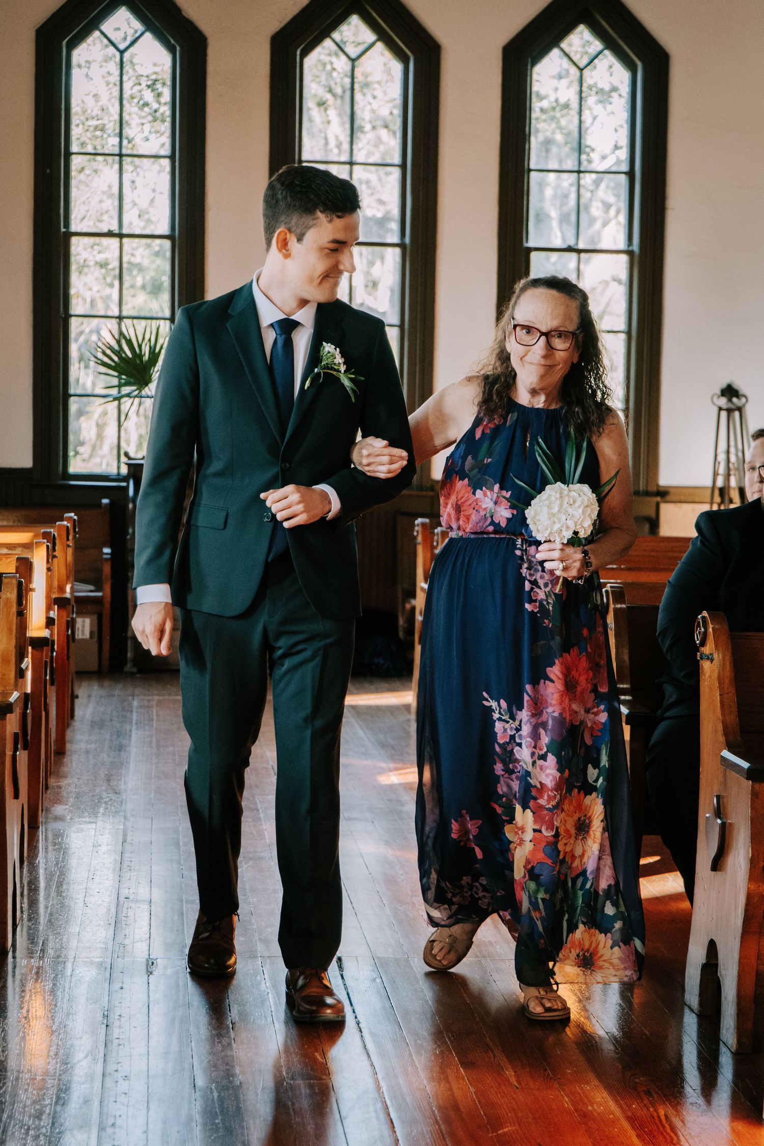 A couple walks down a church aisle with Gothic windows, the woman wearing a navy floral dress and man in dark suit.