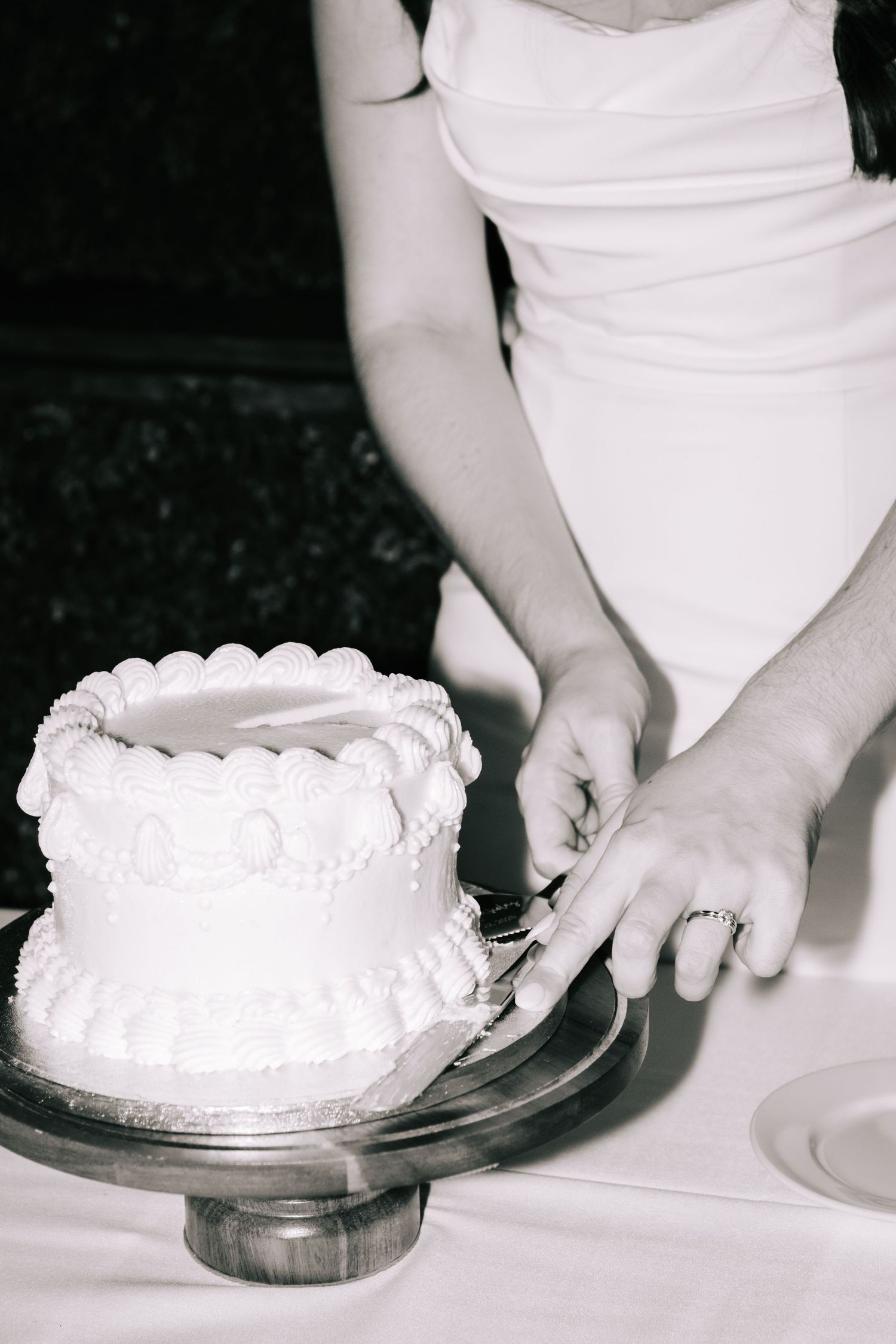 Black and white close-up of white wedding cake being cut on glass serving plate.