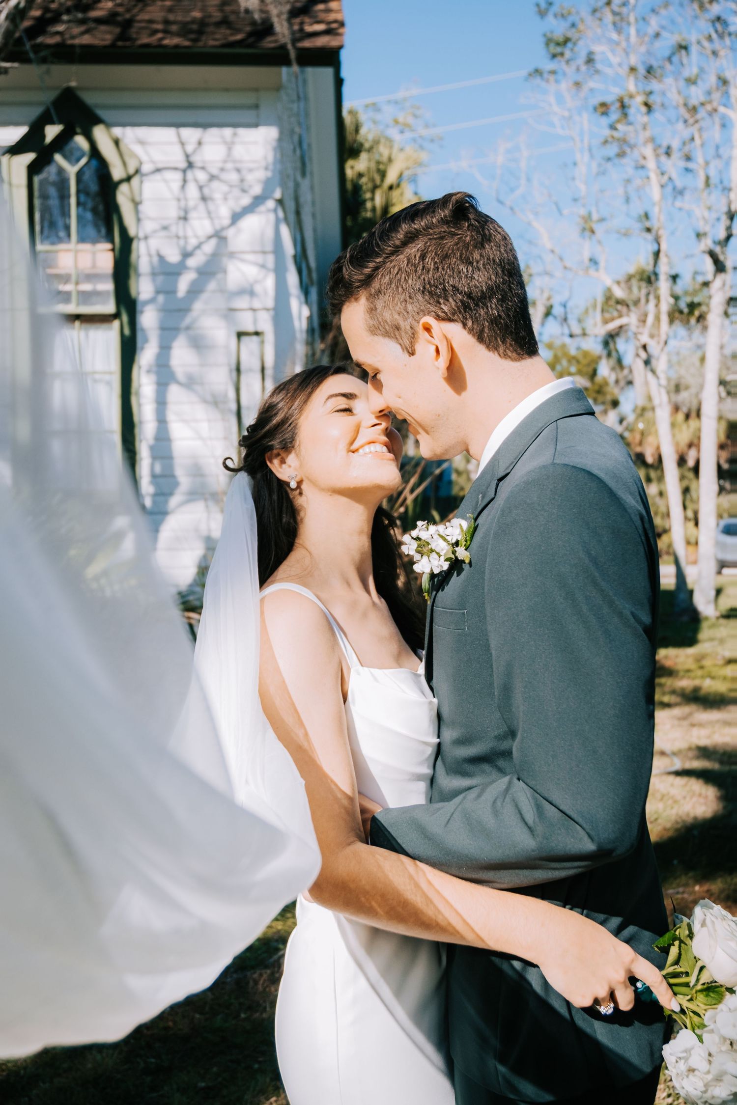 A tender moment between newlyweds as the veil flows in the breeze by a white chapel.