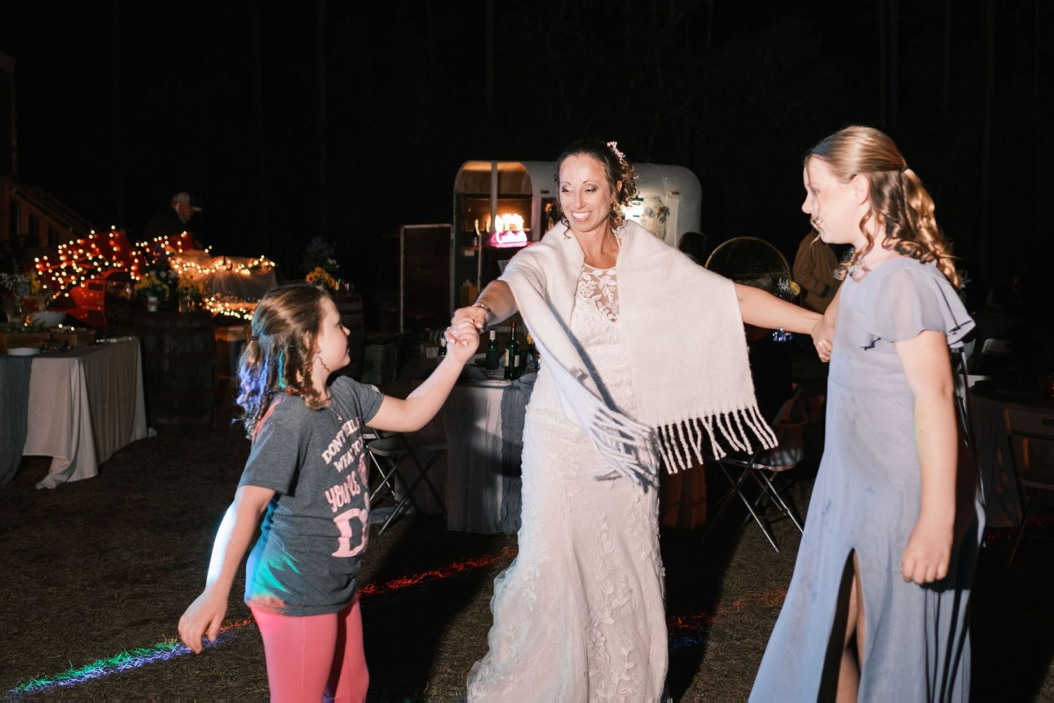 Bride in white lace gown dances joyfully with two young girls at evening wedding reception.
