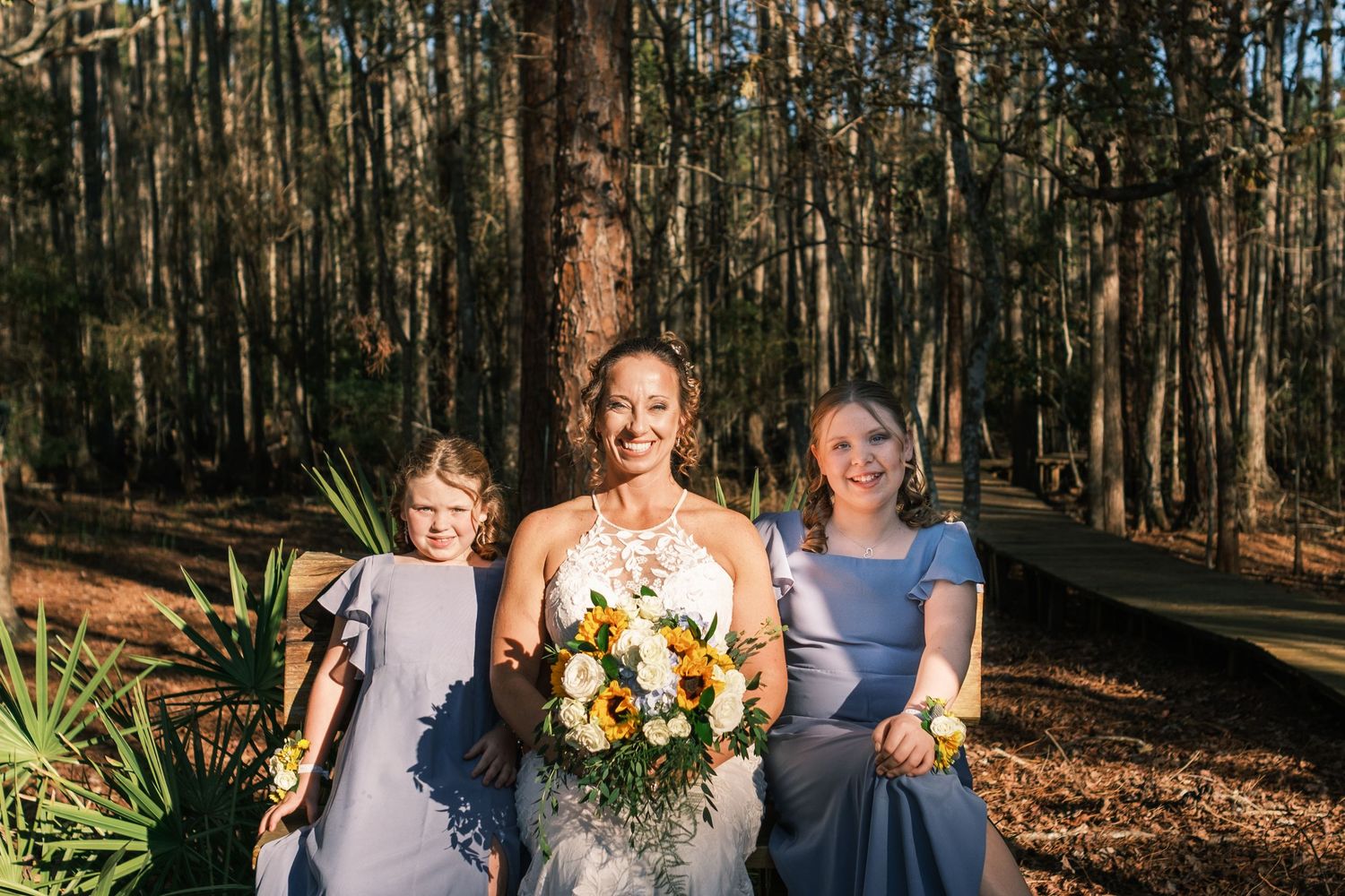 Bride in lace gown holds yellow bouquet flanked by two young bridesmaids in blue dresses in forest.
