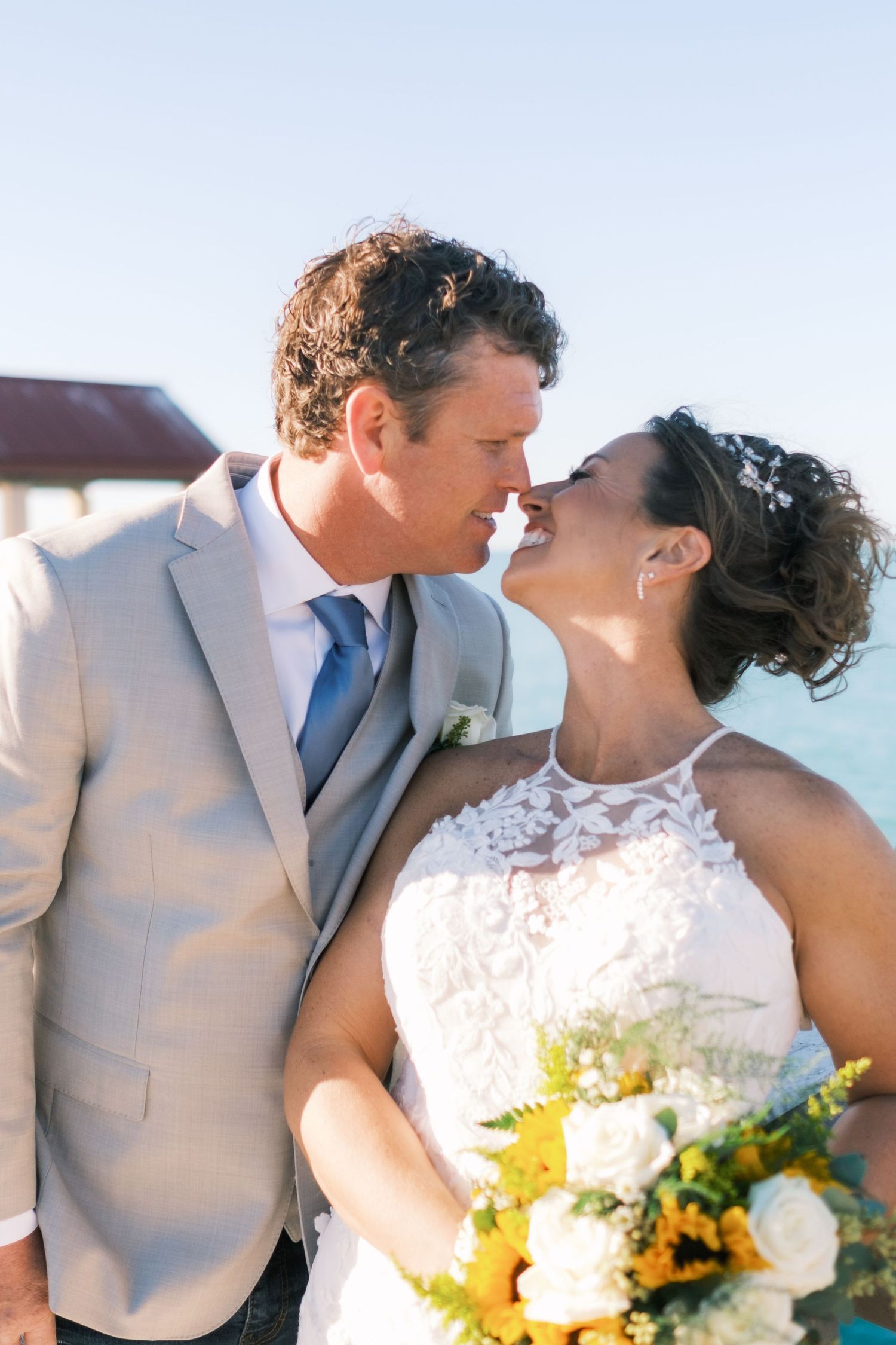 Happy wedding couple almost kissing outdoors, groom in tan suit with blue tie, bride holding yellow and white bouquet.