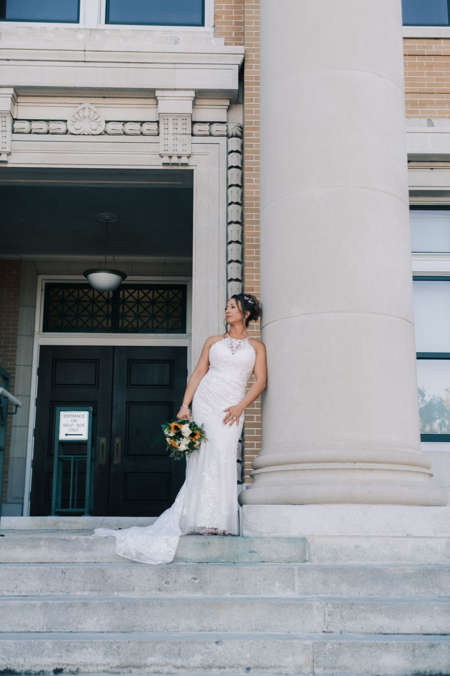 Elegant bride in lace gown holds bouquet beside a large column on building steps, gazing upward.