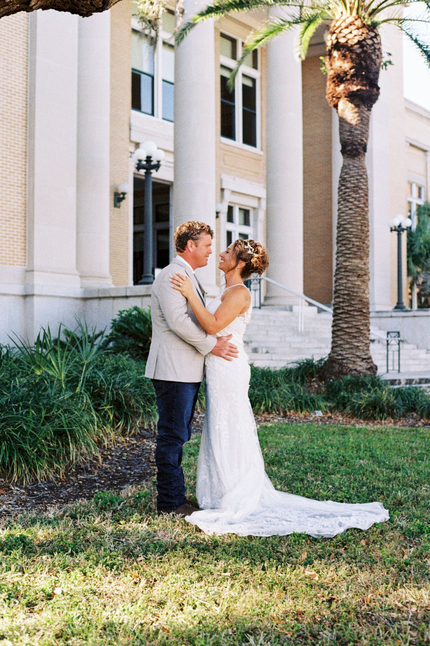 Bride and groom kissing in front of building with palm tree at outdoor wedding venue.