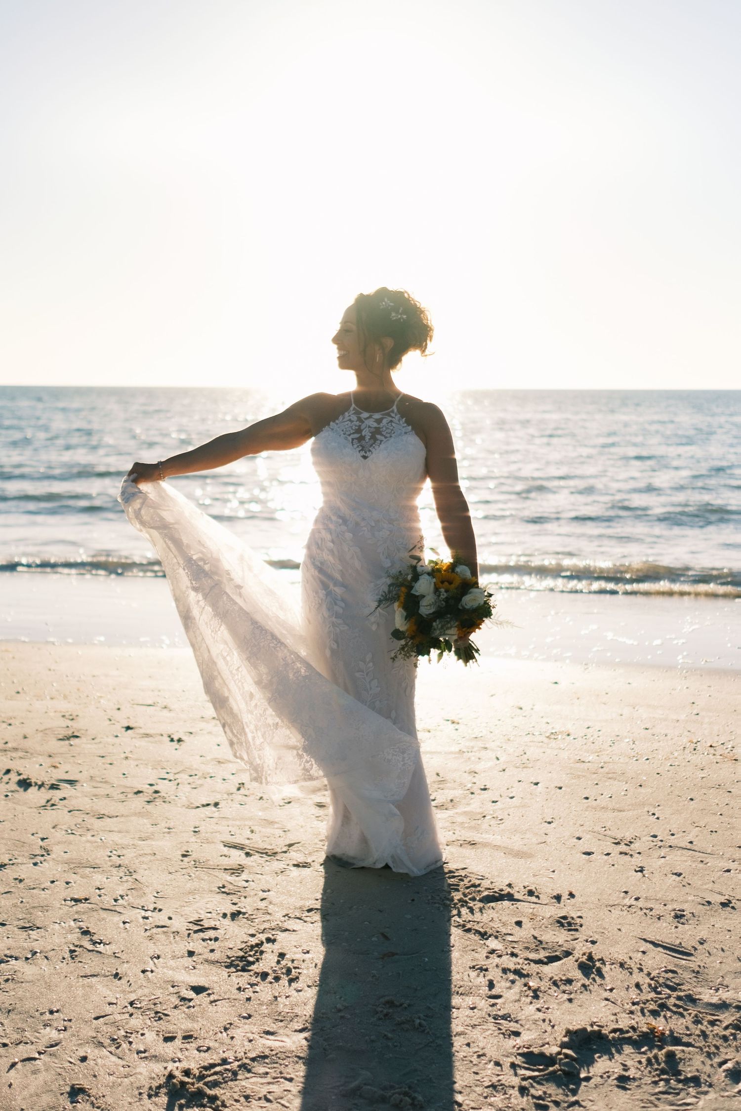 Bride in white lace gown holds bouquet and veil on sandy beach at golden hour sunset.