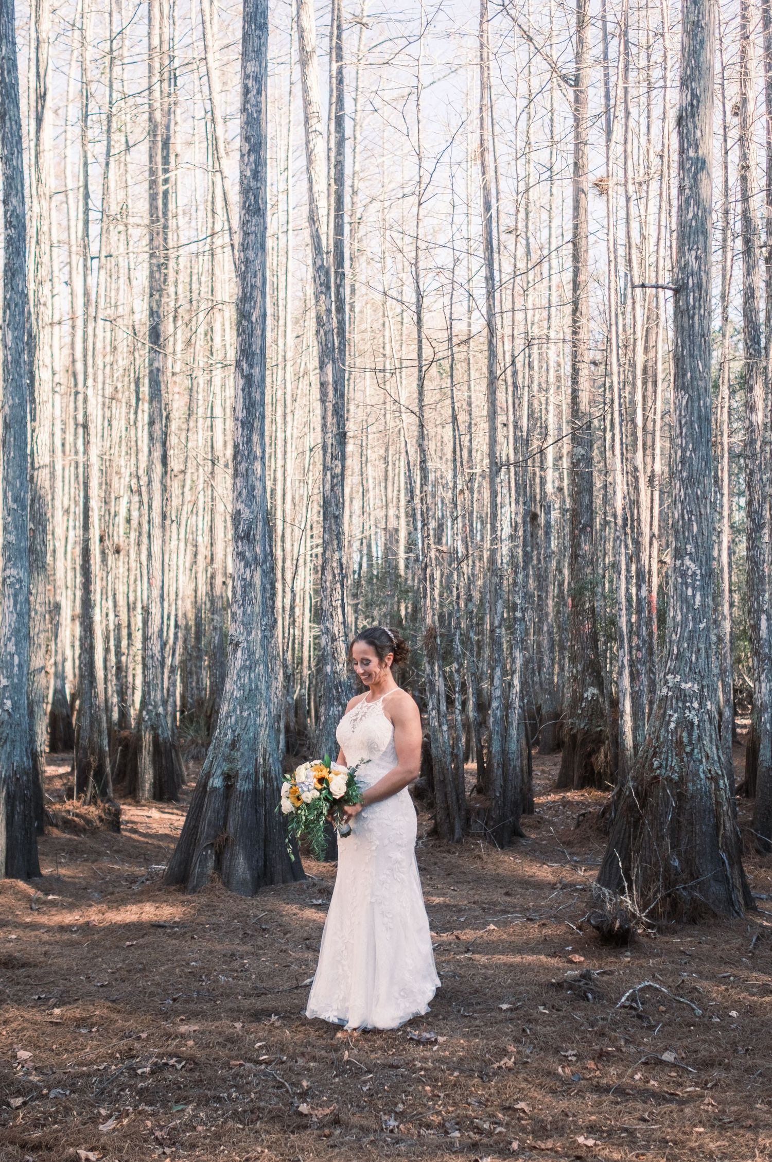 Bride in lace gown holding bouquet among tall bare trees in forest.