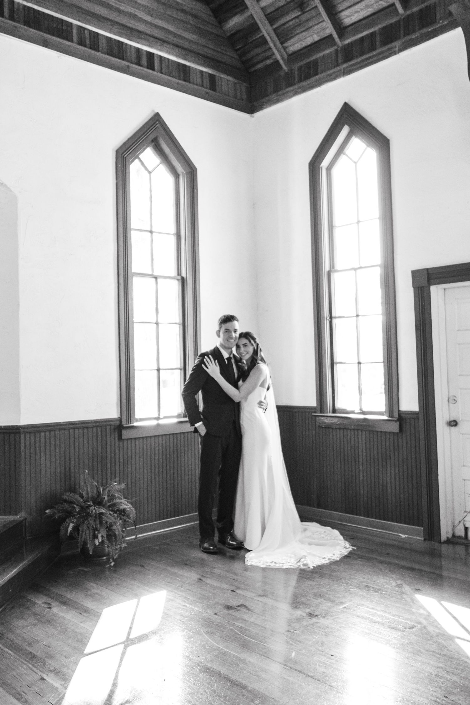 A black and white wedding portrait in a historic chapel with tall gothic windows.
