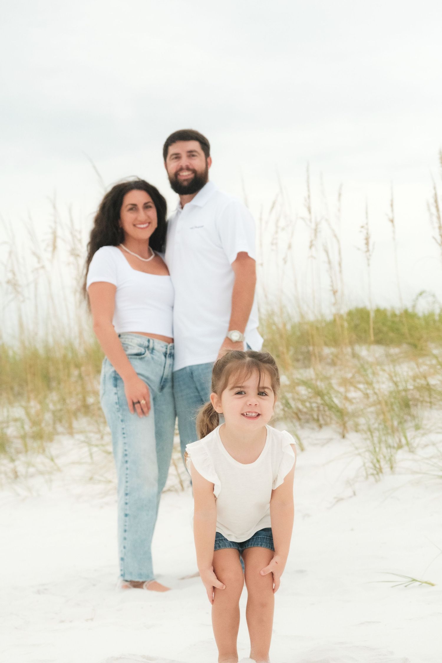 Family wearing white and denim poses together on a sandy beach with sea oats in the background.