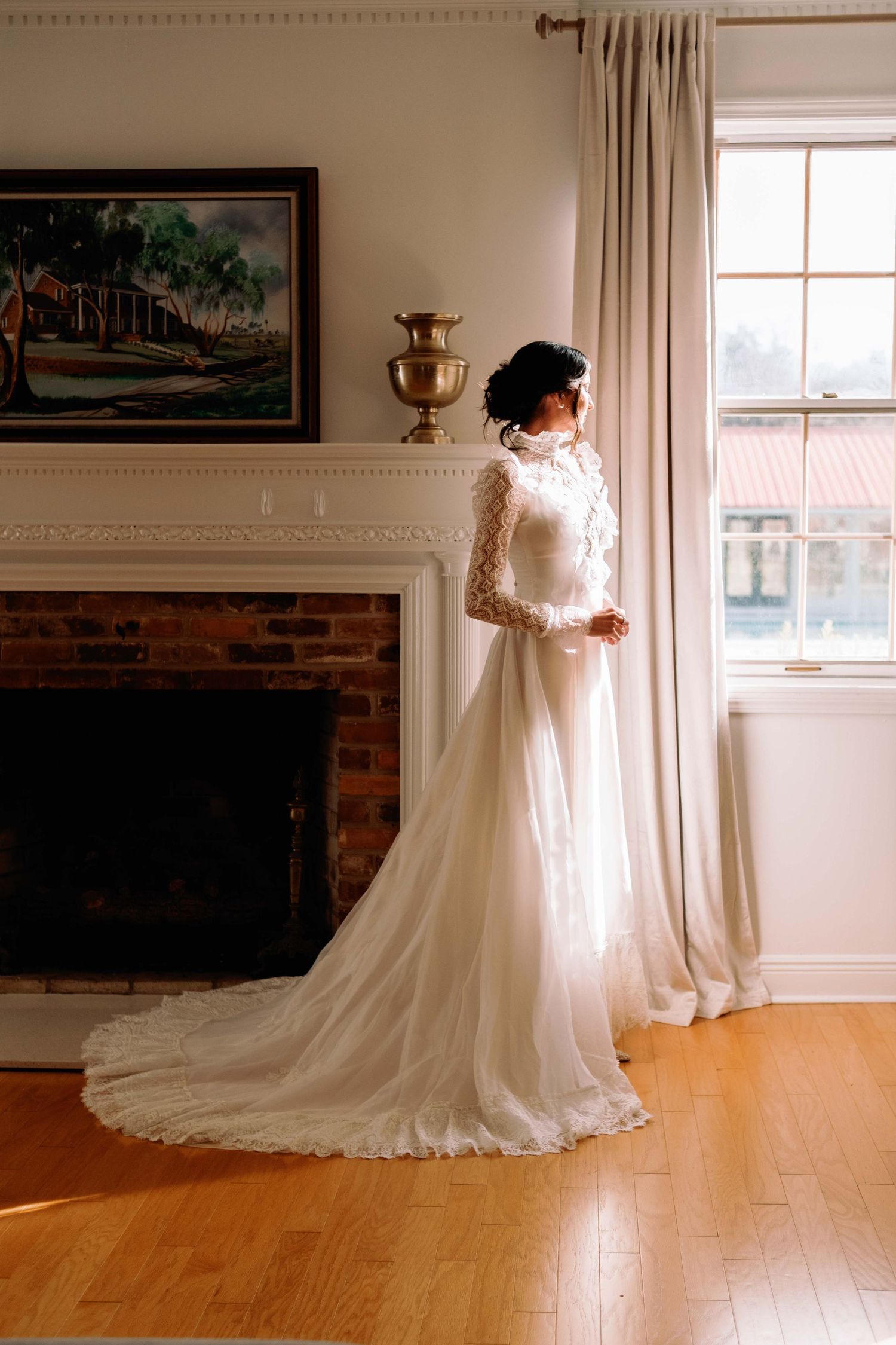 Bride in elegant long-sleeve white lace wedding gown with train stands by sunlit window near brick fireplace.
