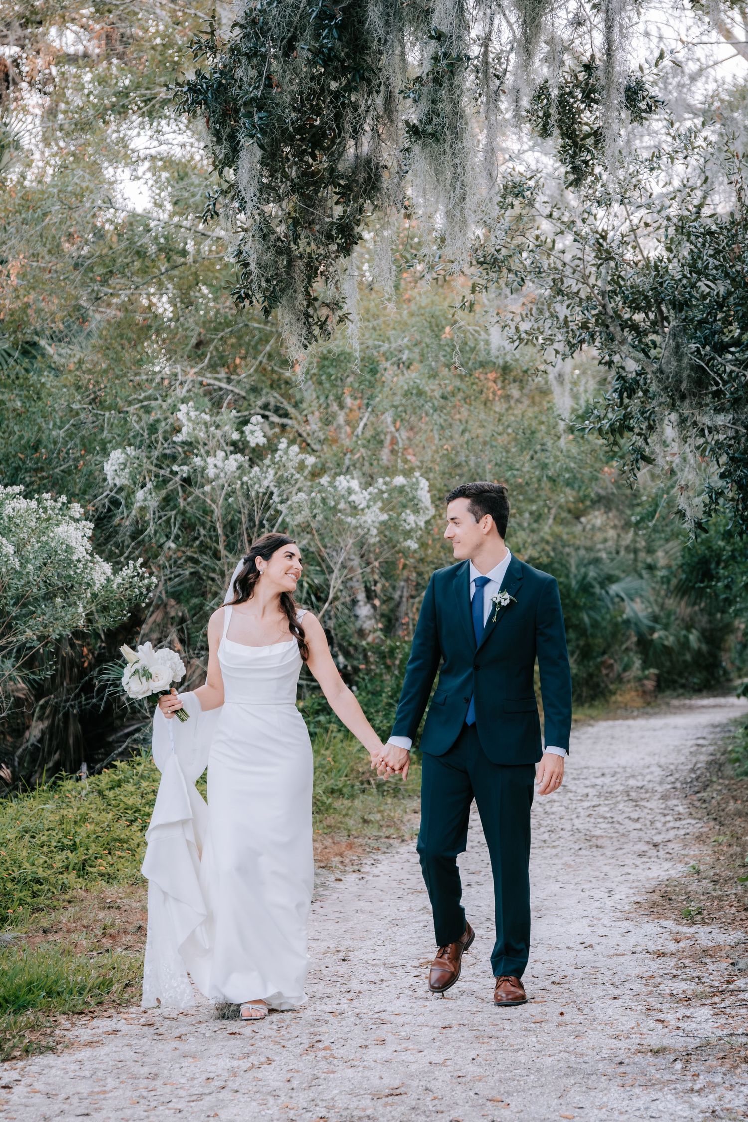 Bride and groom walking together on sandy path surrounded by Spanish moss trees.