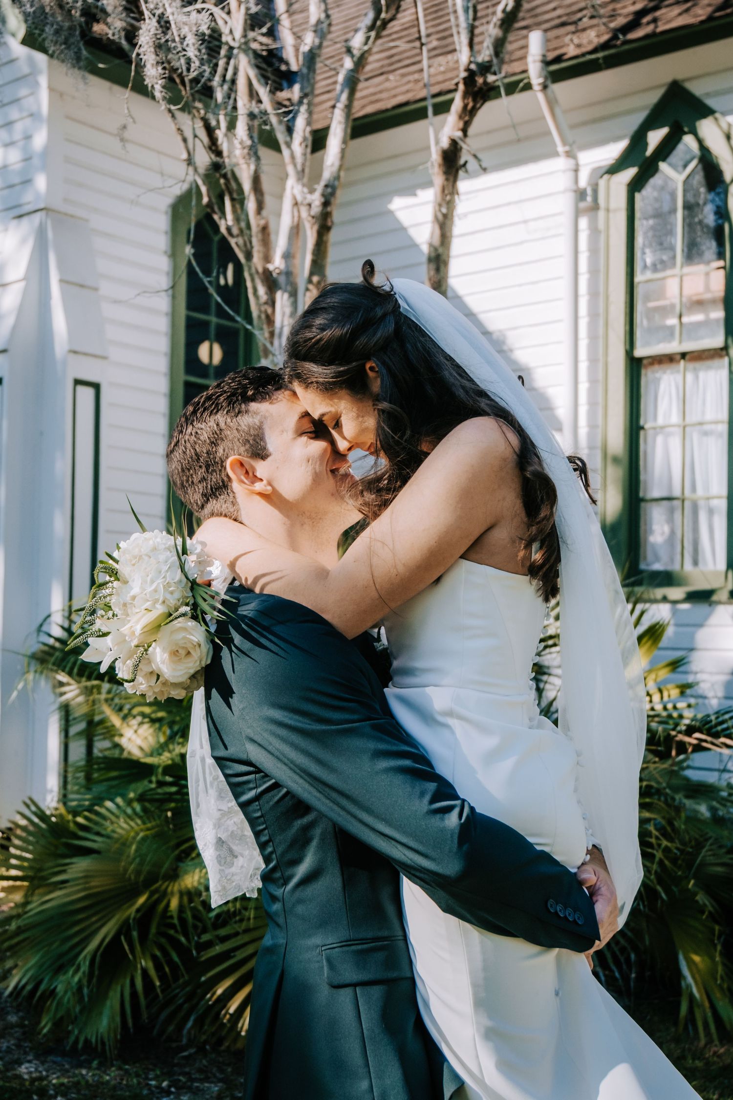 Romantic wedding couple embraces outside a white colonial-style building with palm trees.