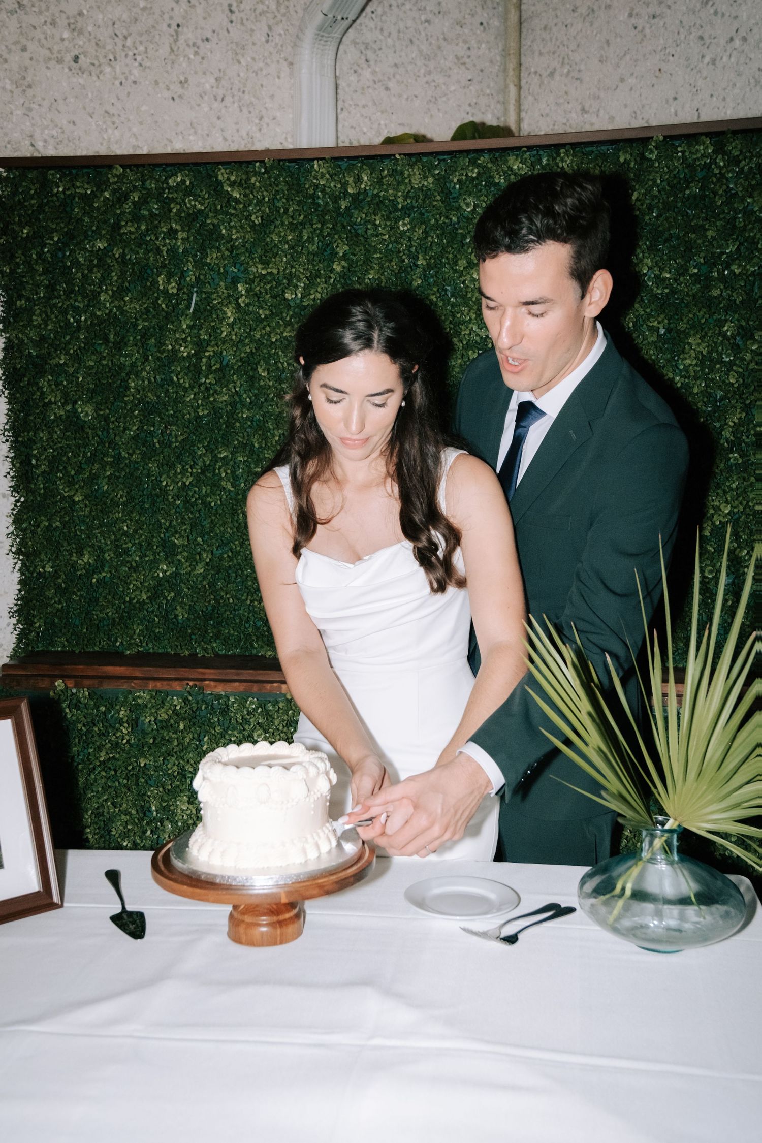 A couple cuts their white wedding cake together at their reception in front of a green hedge wall.