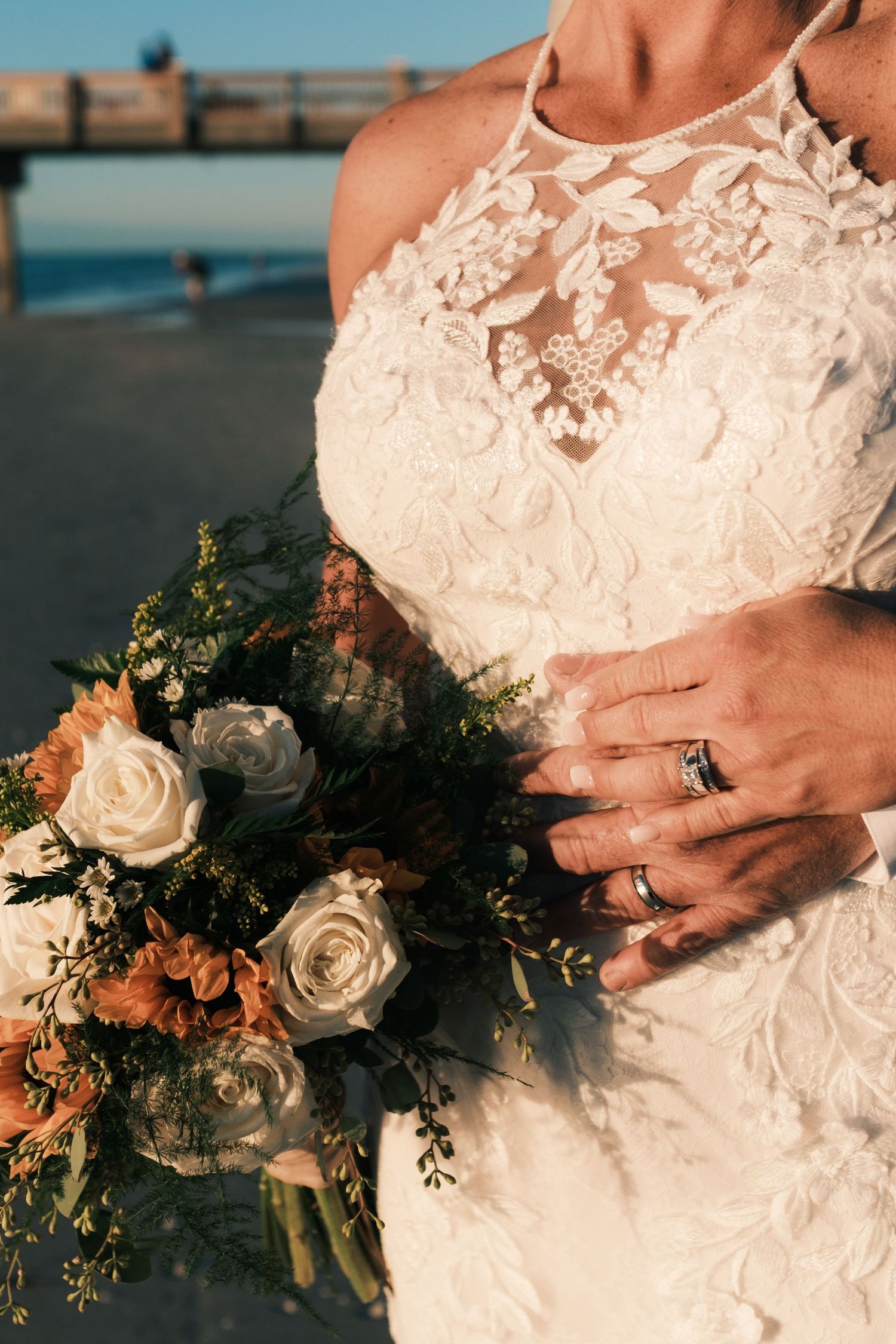 Bride in lace halter wedding gown holds rustic bouquet of white roses and orange flowers at beach sunset.