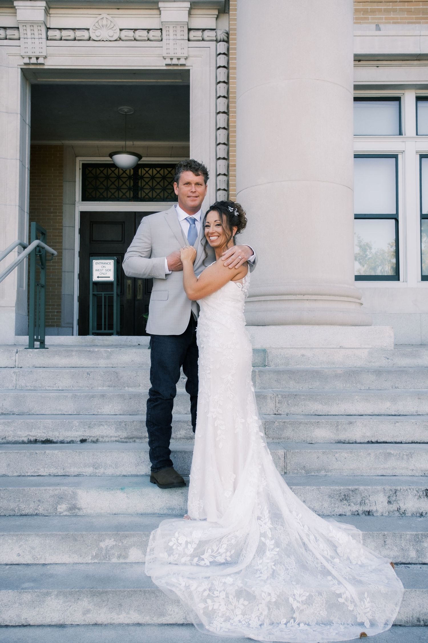 Bride in lace gown with train and groom in gray suit posing on steps of classic stone building.