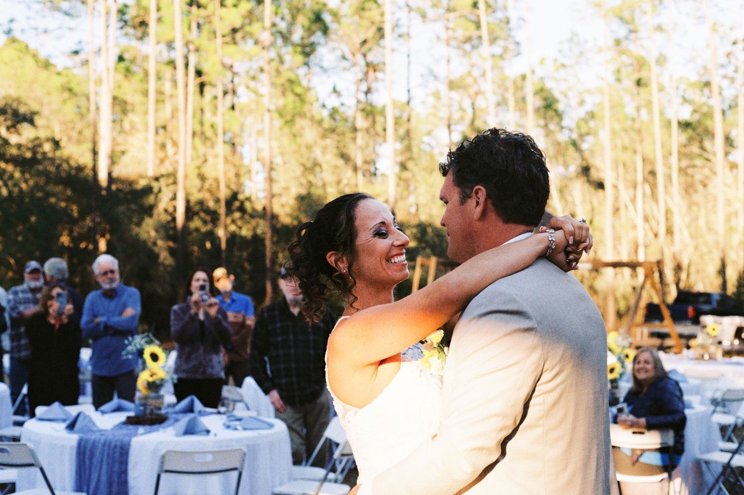 Smiling bride embraces groom during outdoor first dance as wedding guests applaud in a forest setting.