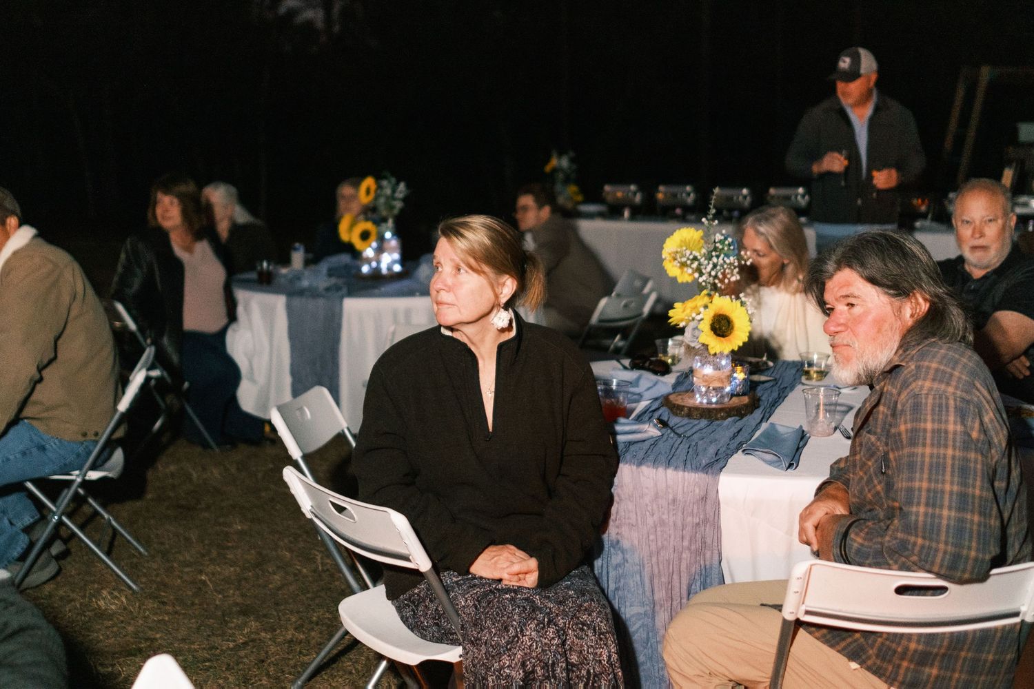 Wedding guests seated at round tables decorated with sunflower centerpieces enjoy an outdoor evening reception.