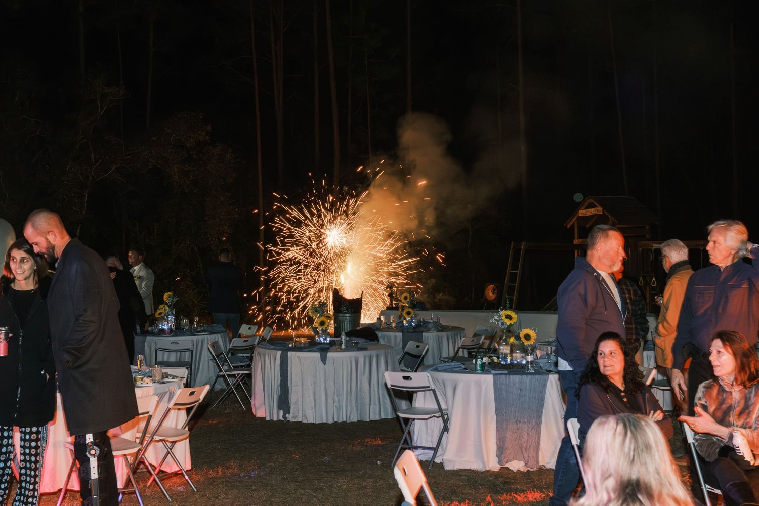 Sparkling fireworks fountain display at outdoor evening wedding reception among seated guests.