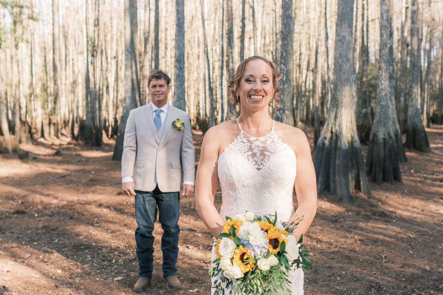 Bride in lace gown holds sunflower bouquet while groom stands behind her in cypress tree forest.