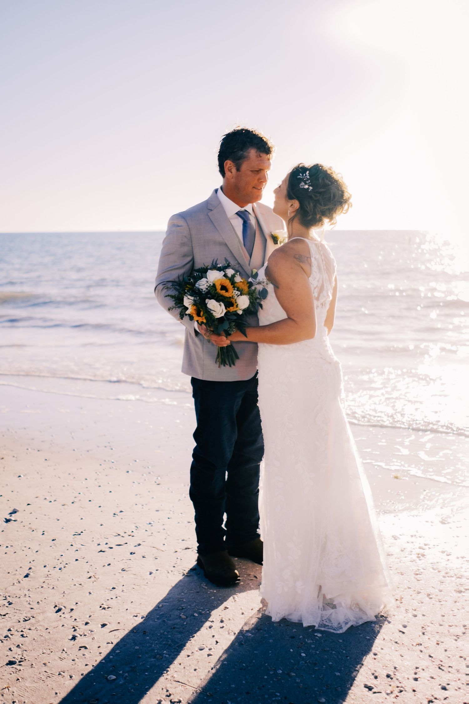 Bride and groom stand facing each other on a sunny beach, sharing a tender moment with a sunflower bouquet.