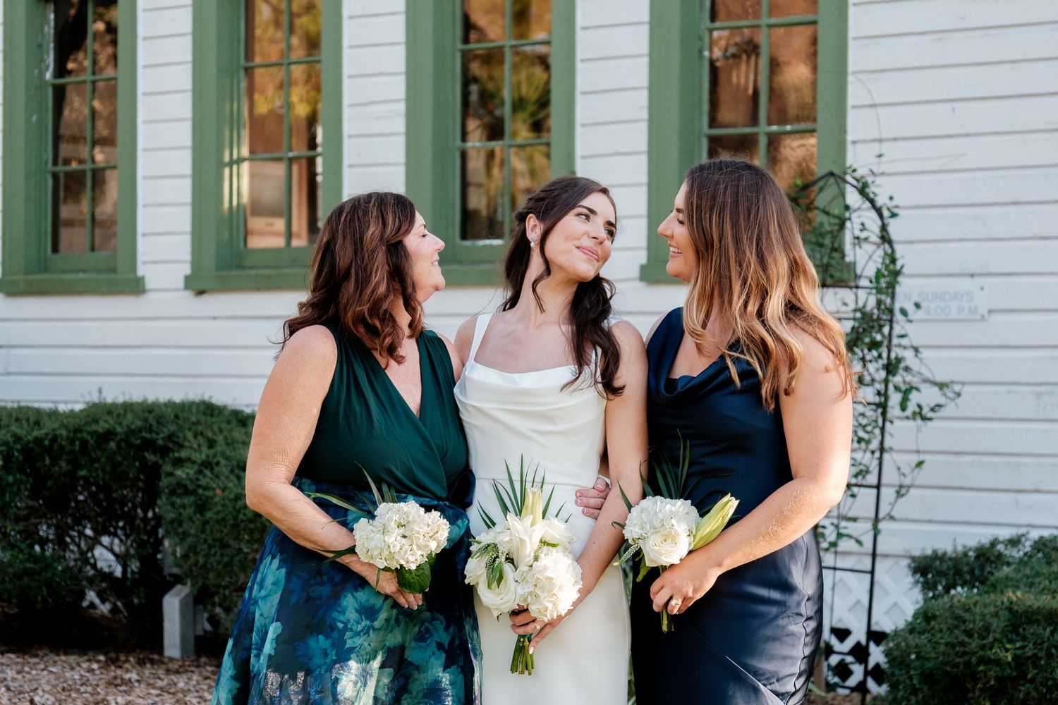 Three women in elegant dresses pose with bouquets against a white building with green windows.