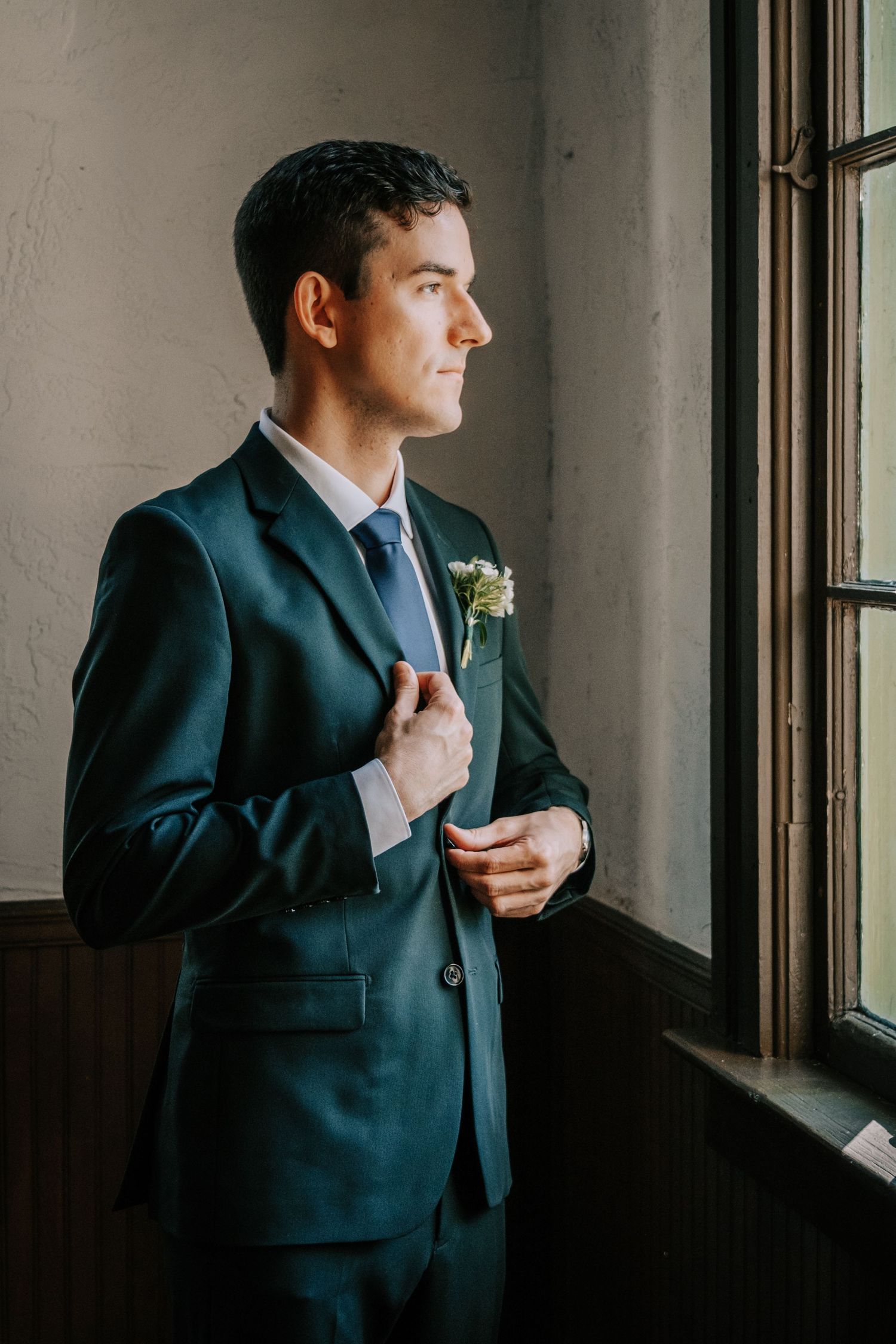Groom in dark suit adjusts tie while looking pensively out window.