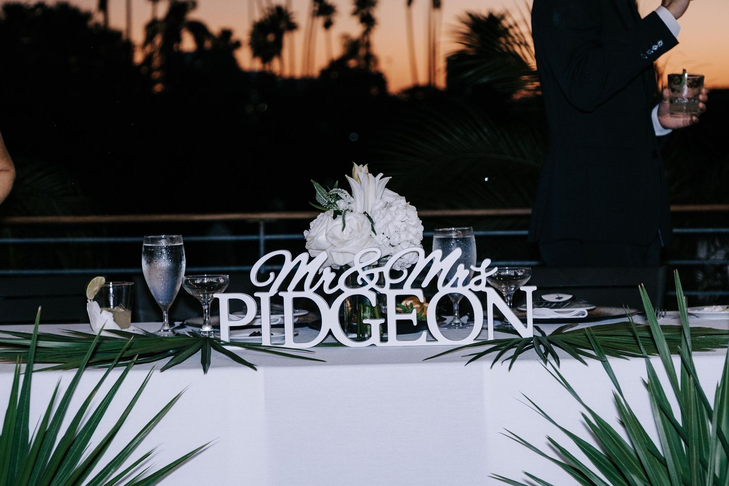 Wedding reception table setting outdoors with palm trees and string lights visible in the evening sky.