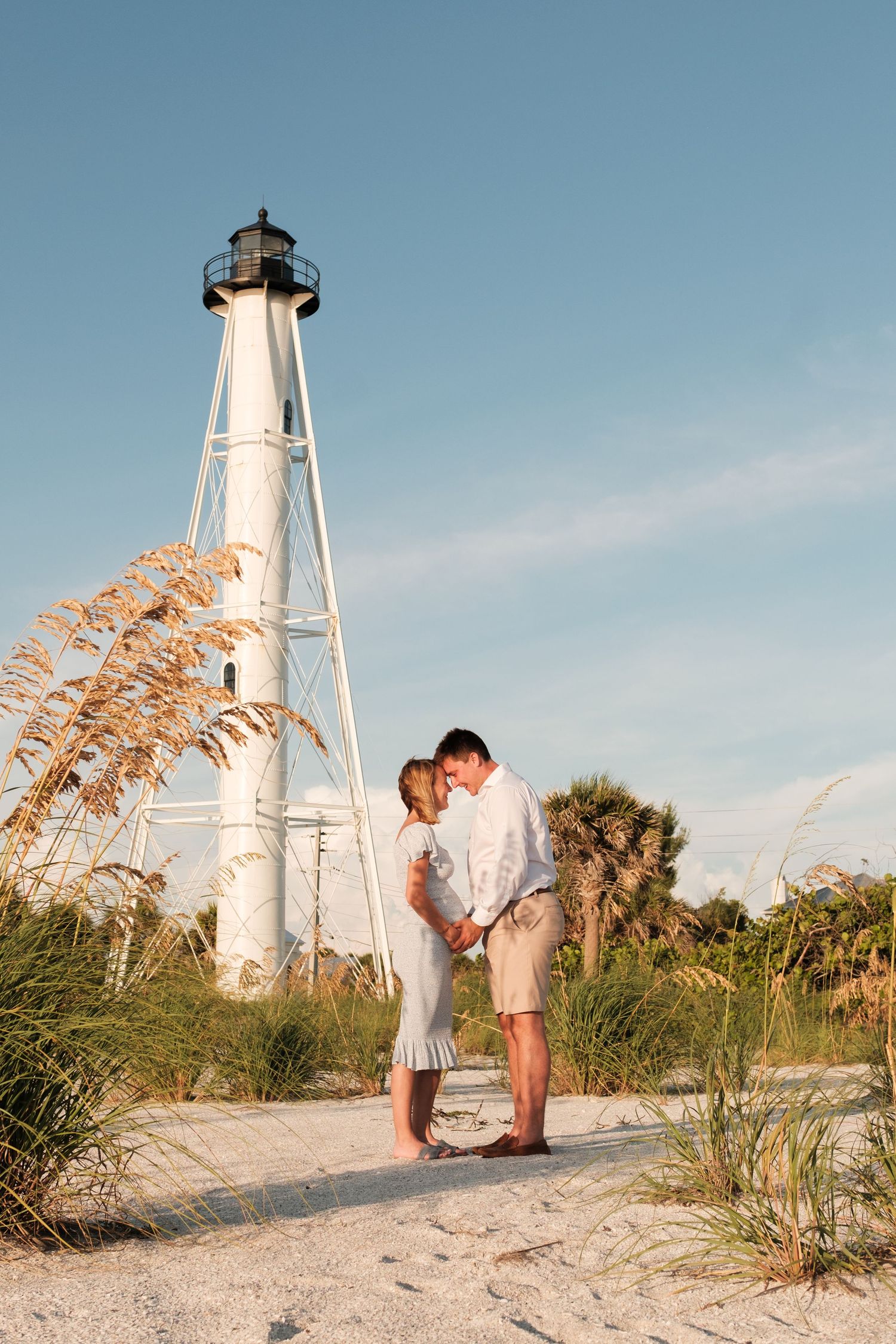 A romantic couple embraces near a white lighthouse on a sandy beach with sea oats and palm trees at sunset.
