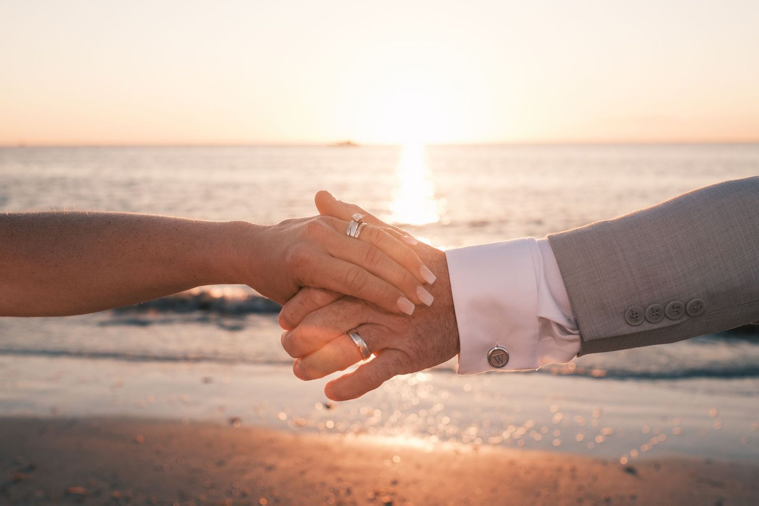 Close-up of bride and groom holding hands at sunset on the beach, wedding rings visible.