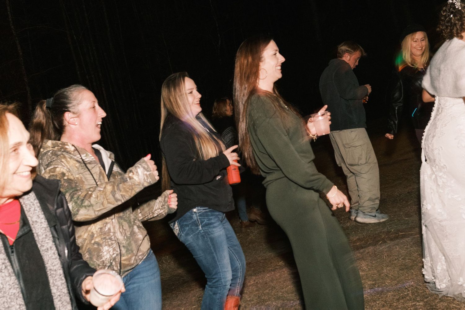 Group of women laugh and dance together at an outdoor nighttime wedding reception celebration.
