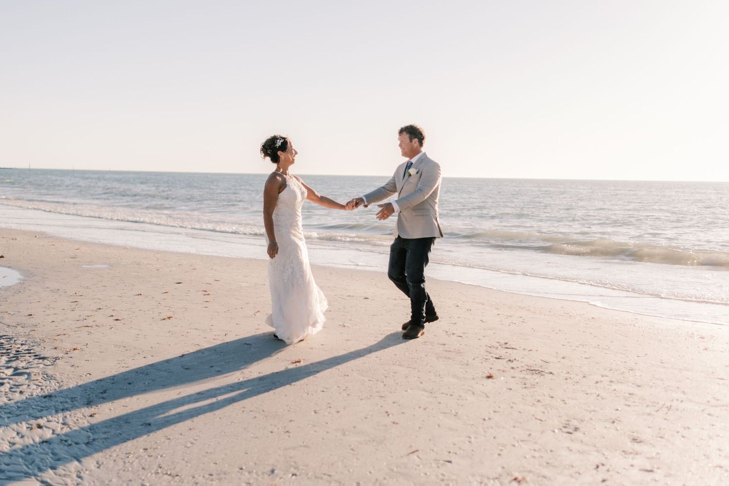 Bride and groom dance and laugh together playfully on a sandy beach during golden hour.