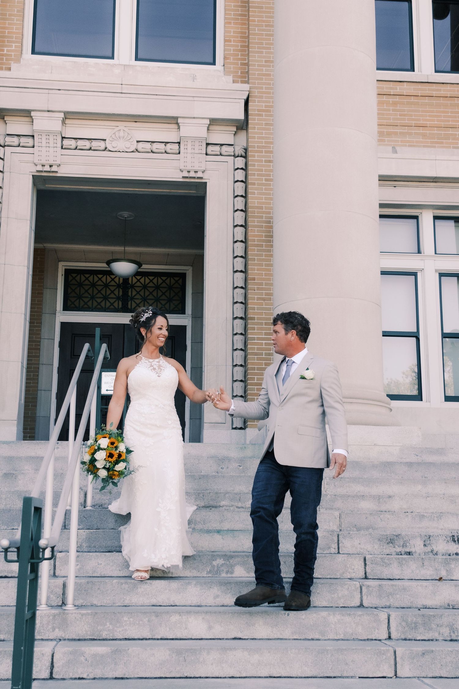 Newlyweds hold hands descending stone steps outside a grand building on their wedding day.