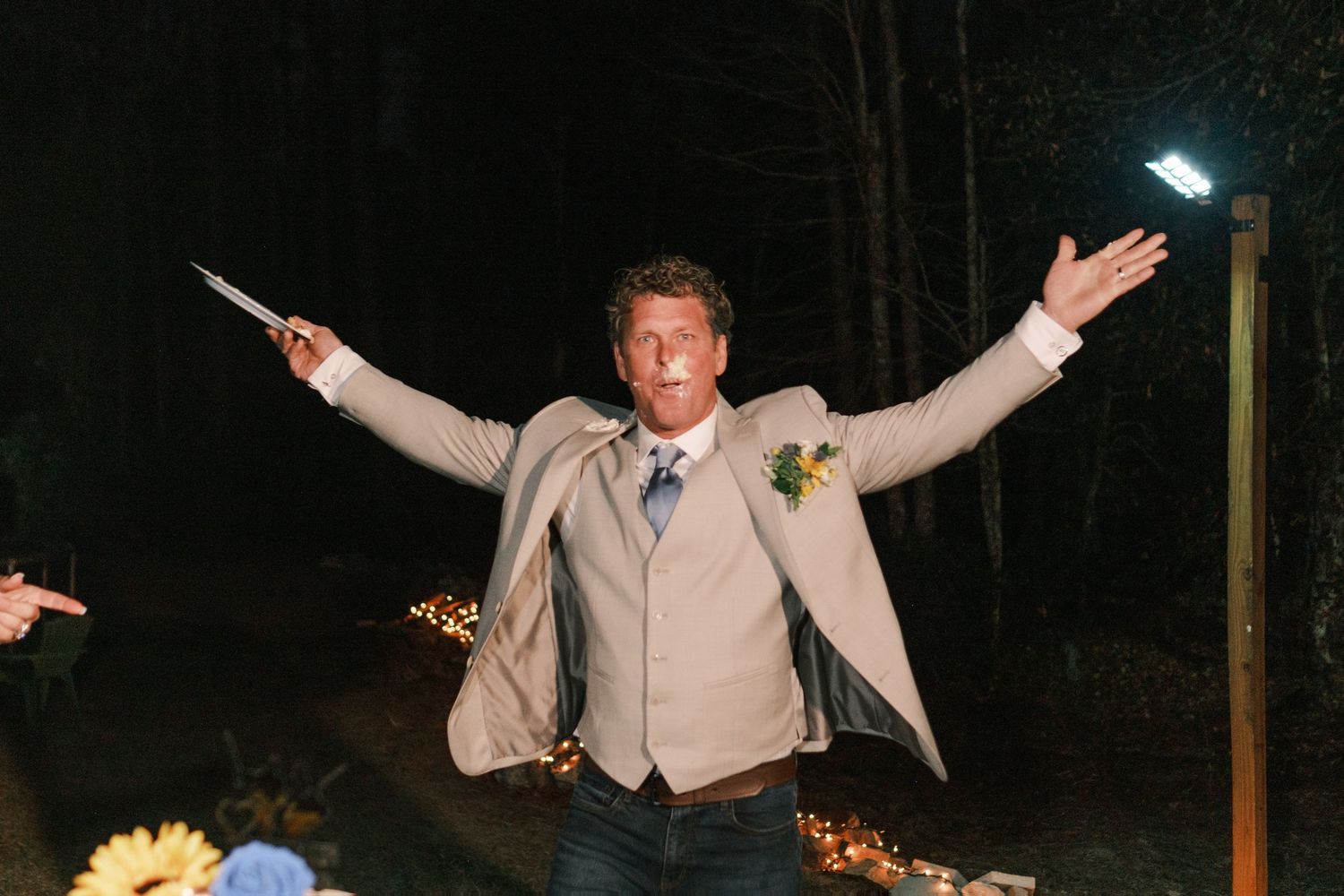 Groom in tan blazer and jeans celebrates outdoors at night with arms outstretched at rustic wedding reception.