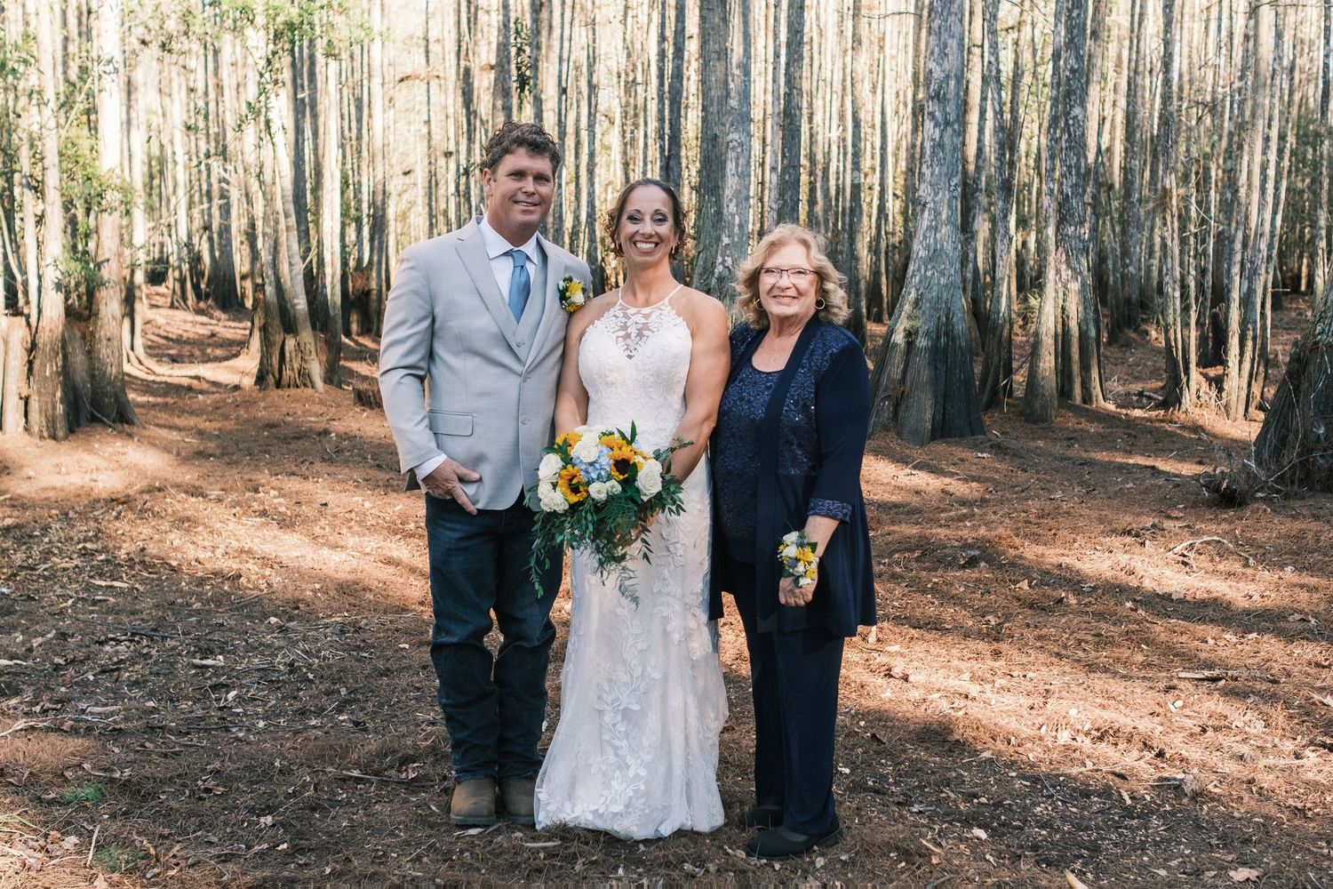 Bride and groom pose with wedding officiant in lush pine forest after outdoor ceremony.