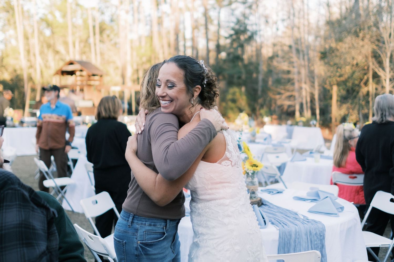 Smiling bride in lace gown embraces a guest at outdoor wedding reception with white tables in background.