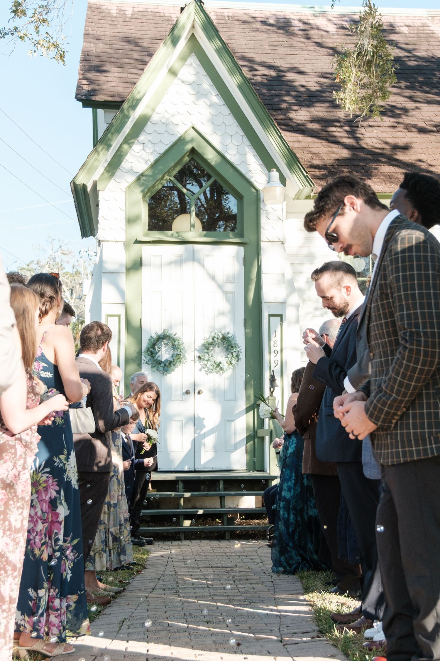 Wedding guests gather outside a charming chapel with a green pointed doorway and wreaths.