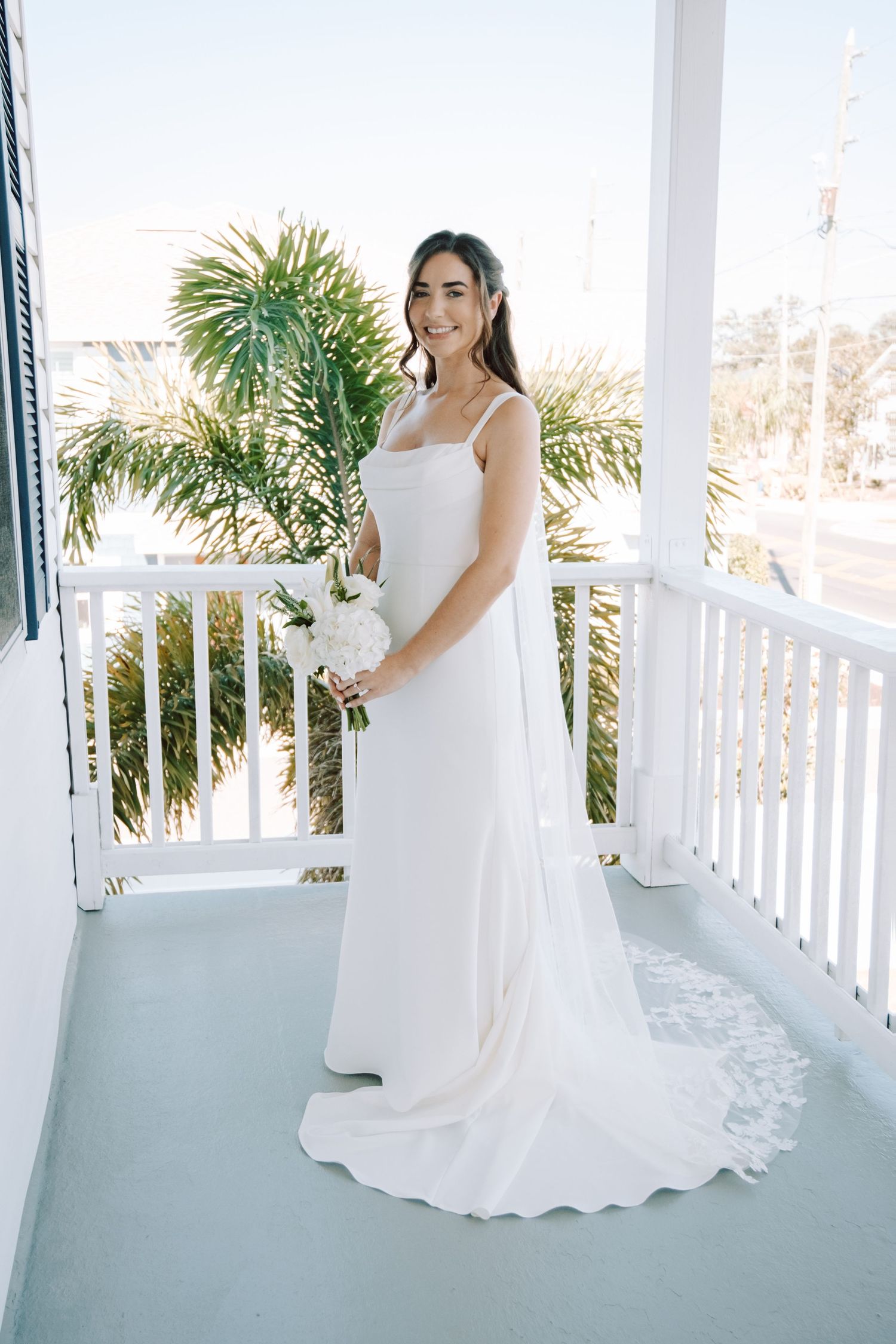 Elegant white wedding dress photographed on a coastal porch with palm trees and white railings in the background.