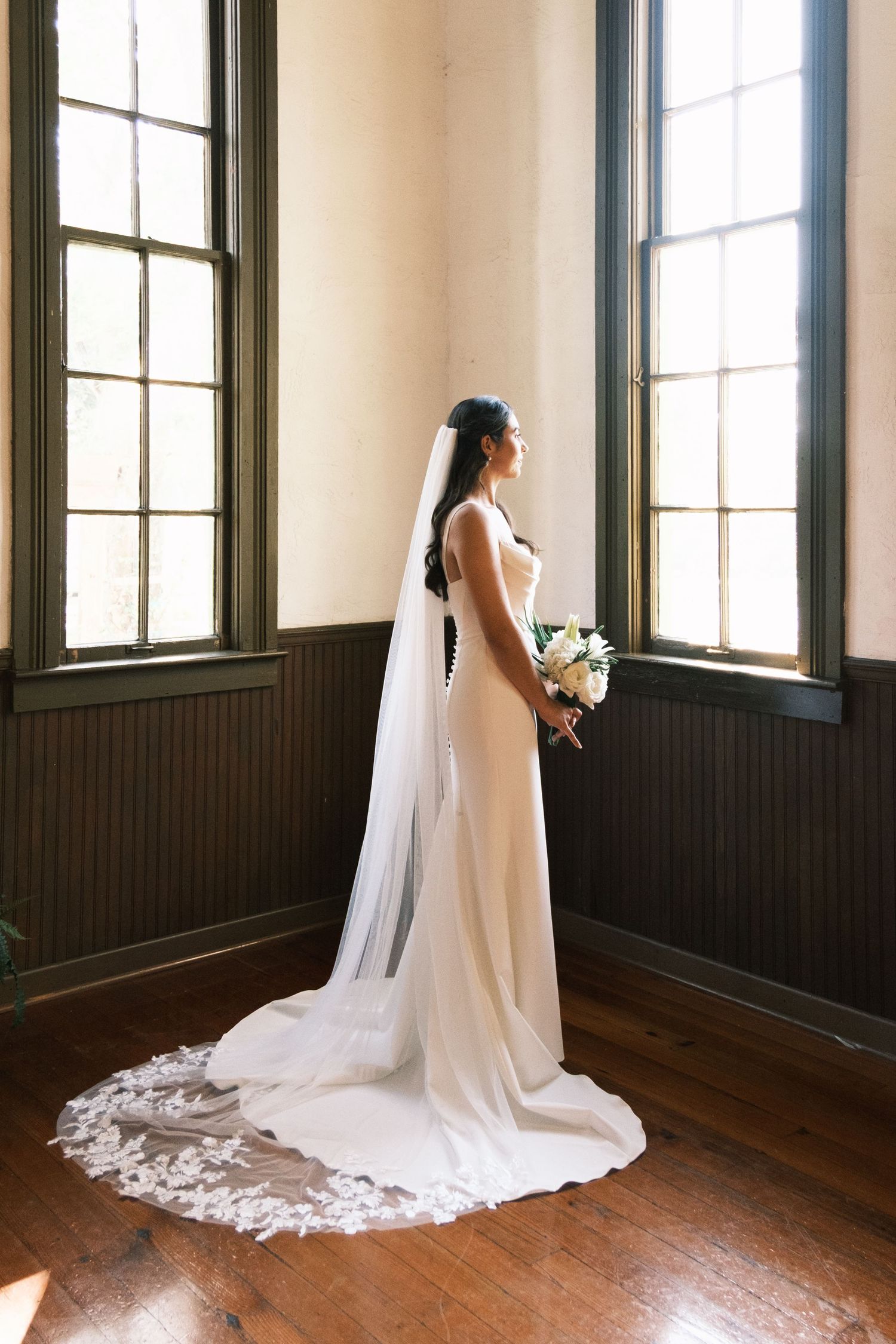 A bride in a flowing white wedding gown and veil stands in a sunlit room with dark wooden floors and tall windows.