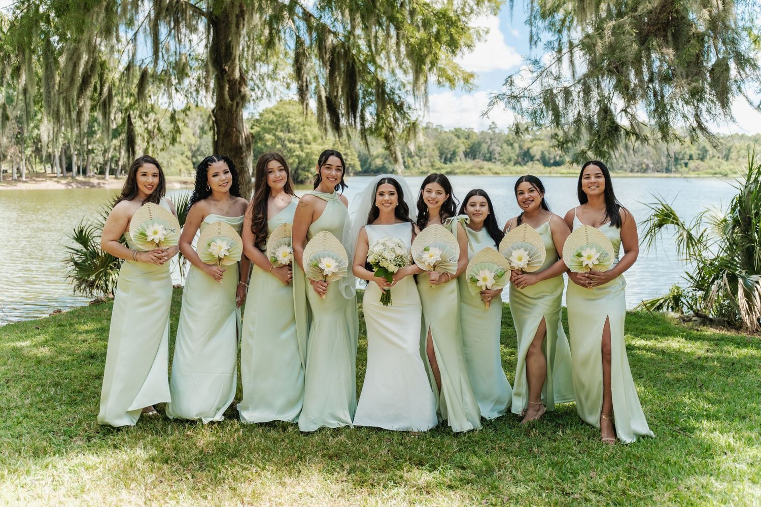 Row of bridesmaids in light-colored dresses standing outdoors under willow trees.