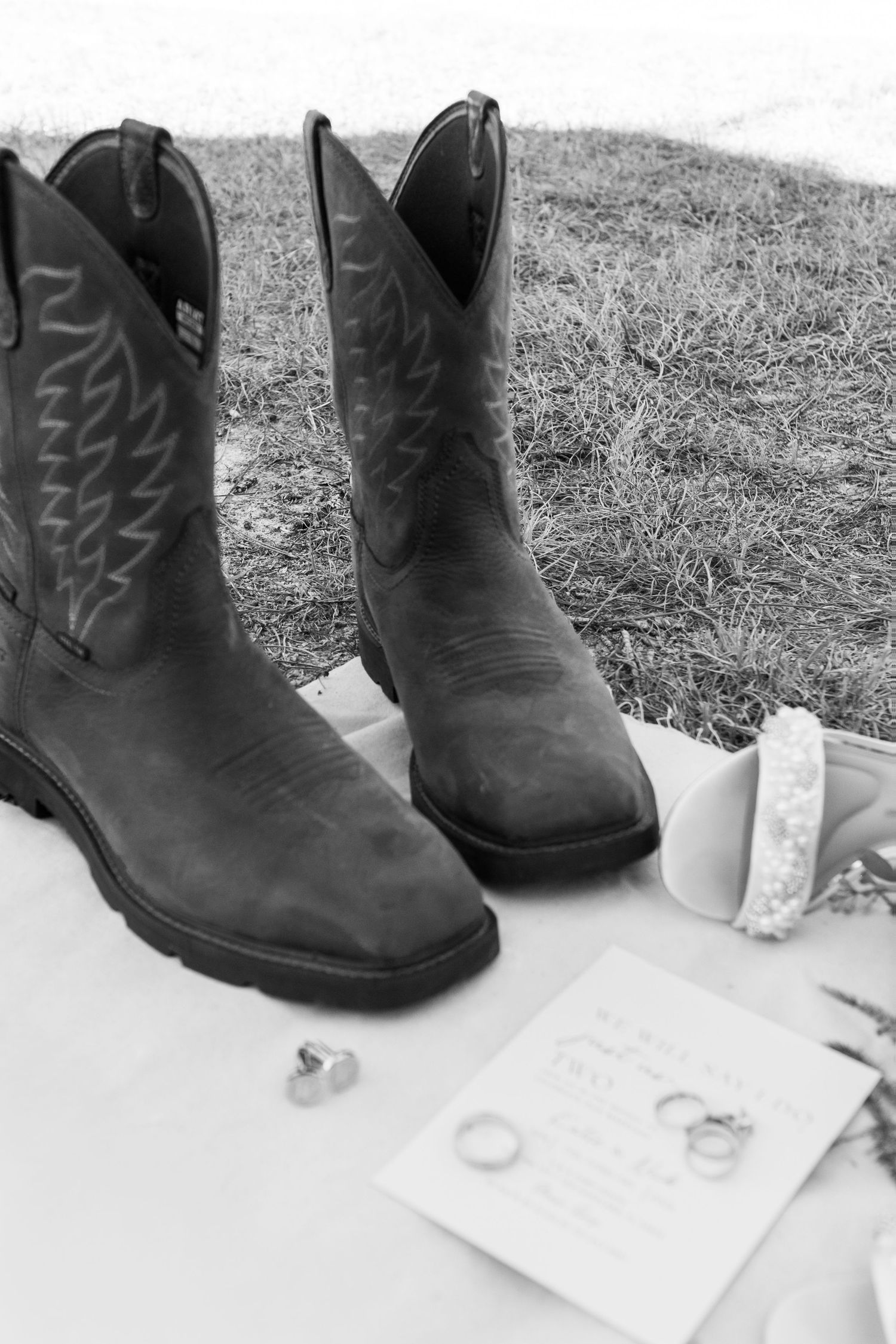 Black and white flat lay of cowboy boots, wedding rings, and invitation on white fabric.