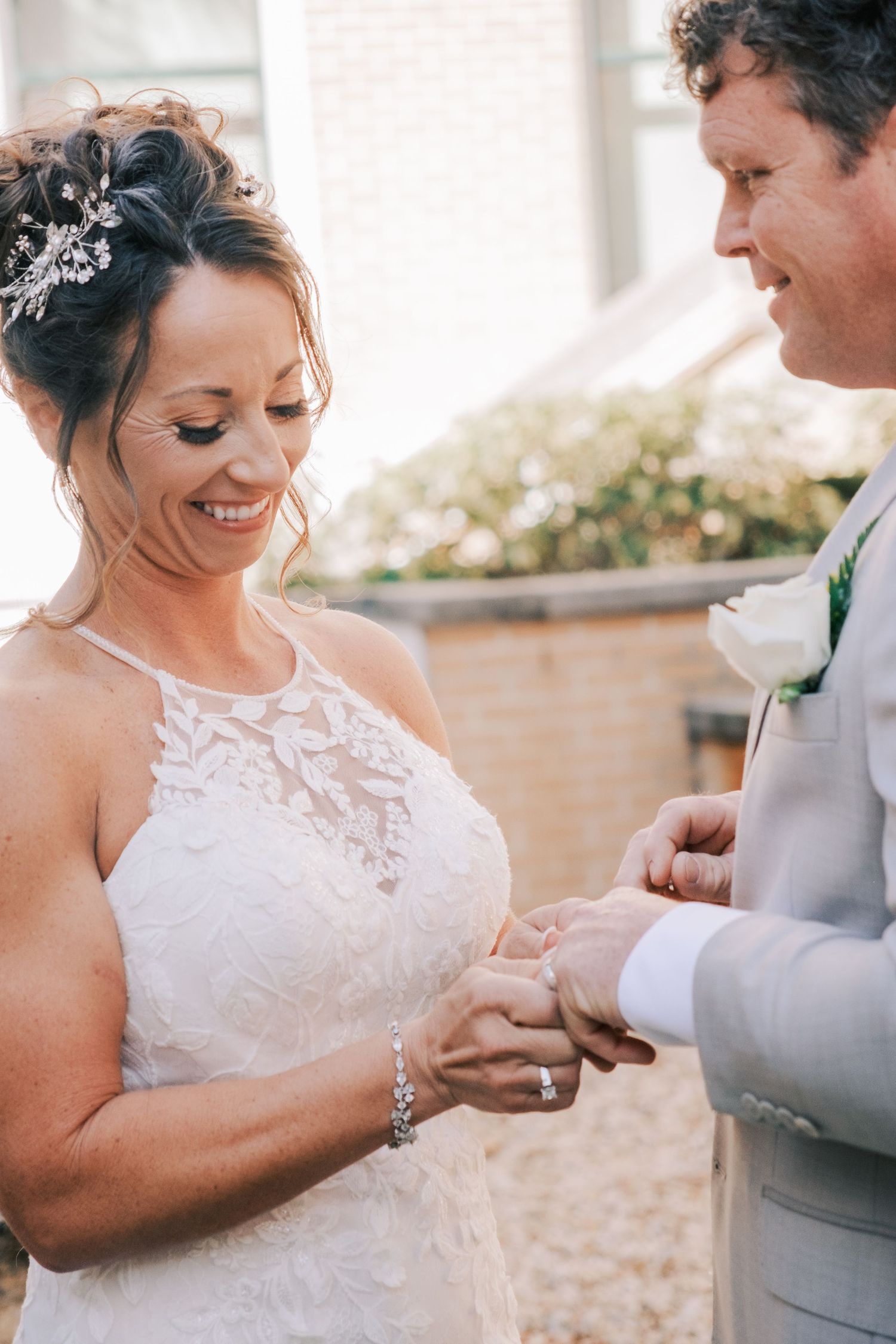 Bride smiles as groom places wedding ring on her finger during outdoor ceremony.