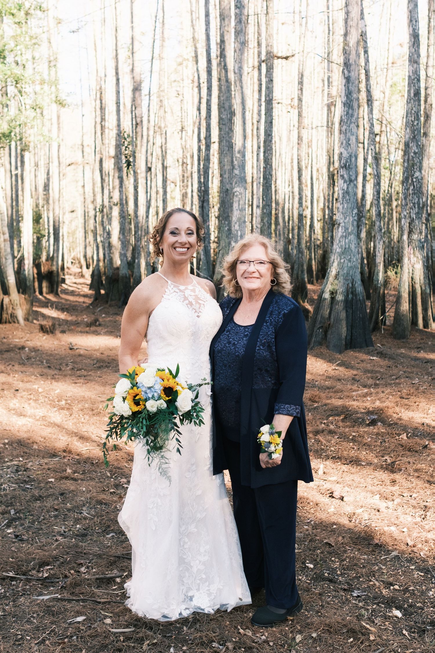 Smiling bride holding a yellow and white bouquet poses with an older woman in navy attire in a pine forest.